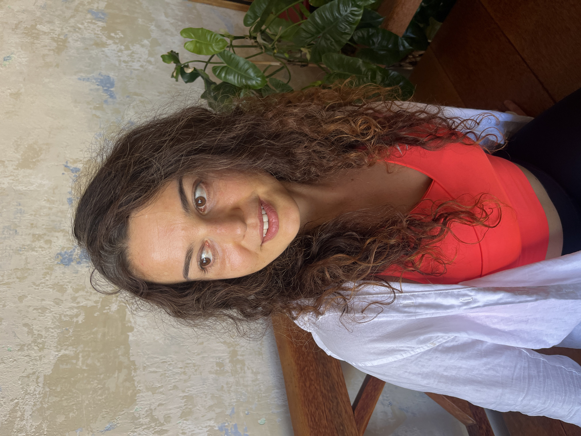 A woman with long, curly brown hair, wearing a white unbuttoned shirt over a bright coral tank top, sits on a wooden bench. She is looking to her left, with a slight smile. Her skin appears tanned and slightly flushed. Behind her, a textured, distressed wall in muted tones of beige, white, and blue is visible. To the right of the woman, a large, leafy green plant stands in a pot, its leaves reaching upwards. The setting appears to be indoors, with wooden surfaces suggesting furniture or paneling. The location is identified as Phu Thuy Ward, Vietnam.