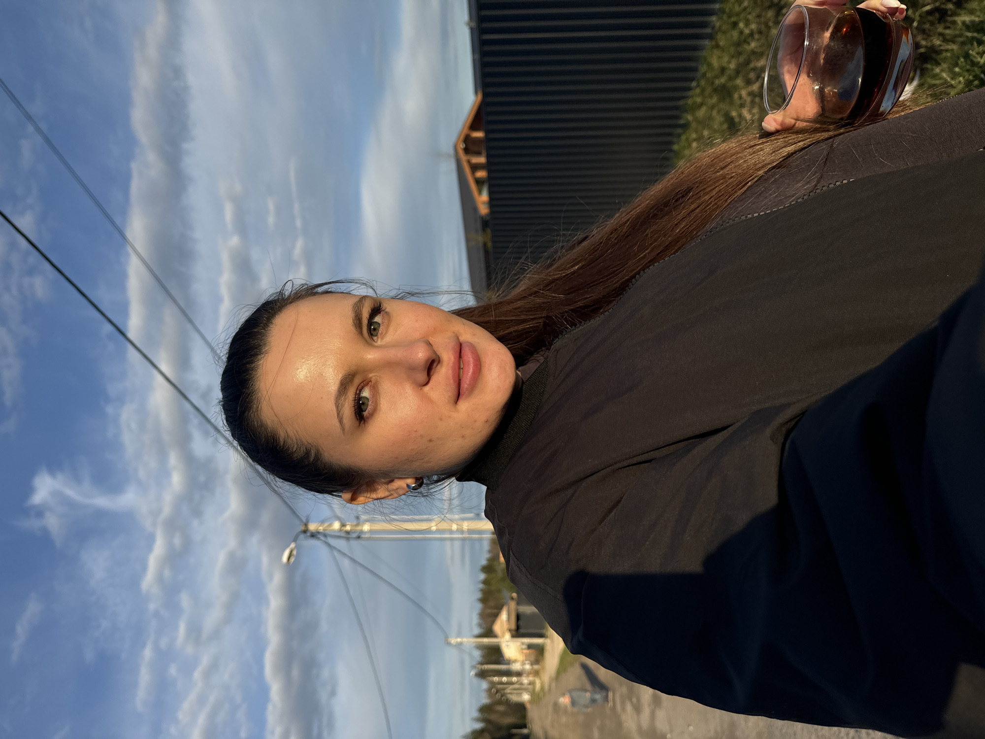 A woman is holding a glass of dark liquid in her right hand, possibly on a street in Литвиново, Russia. She has brown hair pulled back, fair skin, and wears a dark-colored top. Behind her, a street with utility poles and buildings is visible, suggesting an urban or suburban environment. The sky is blue with clouds.