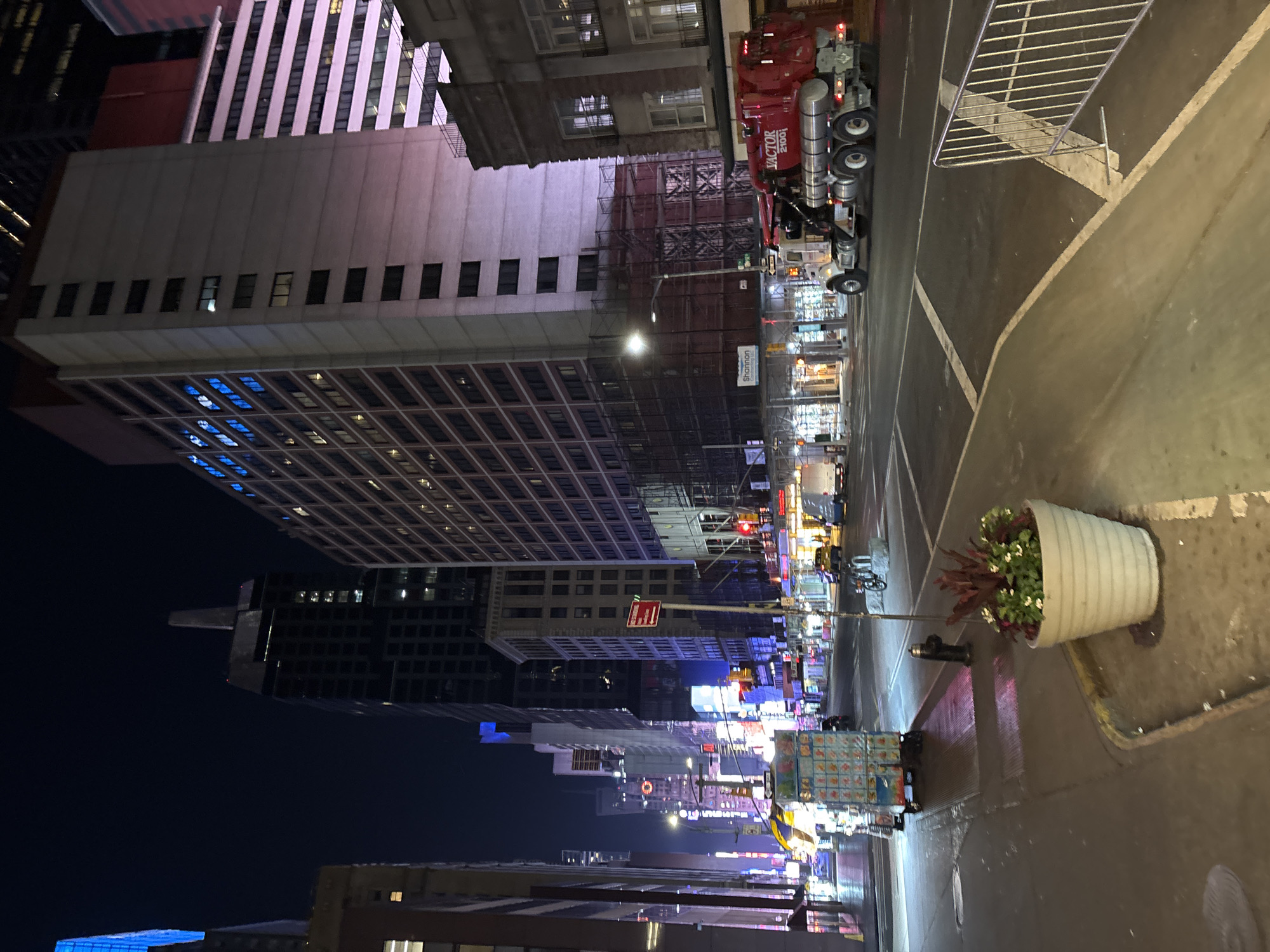 This media file captures a vibrant cityscape at night in New York, United States of America. The scene is dominated by tall, multi-story buildings that line a busy urban street, receding into the distance.
The primary subjects include a prominent red cement mixer truck, clearly visible on the right side of the street, with "VICTOR 2100i" printed on its side. Numerous high-rise buildings frame the street, some featuring illuminated windows with blue reflections, while others display distinct architectural patterns like vertical white stripes or a grid-like façade. Scaffolding is visible on several buildings, suggesting ongoing construction or maintenance.
On the street and sidewalk, a white planter filled with green and red foliage stands near the foreground. A metal barrier or fence is visible on the asphalt, marked with white road lines. Further down the street, streetlights, traffic lights, and a multitude of illuminated commercial signs create a bustling atmosphere. A red "ONE WAY" sign is discernible. A utility cart is also present on the sidewalk.
The activity depicted is the general hustle and bustle of a major city at night, with vehicles on the street and the constant glow of urban life. The time of day is unequivocally night, as indicated by the dark sky and the pervasive artificial illumination from buildings, streetlights, and neon signs. The street appears damp, possibly from recent rain, reflecting the bright lights. The overall mood is energetic and vibrant, typical of a bustling commercial district in a large city.
Visible text includes "VICTOR 2100i" on the cement mixer, a red "ONE WAY" sign, and the partial word "Shannon" on a white sign further down the street.
