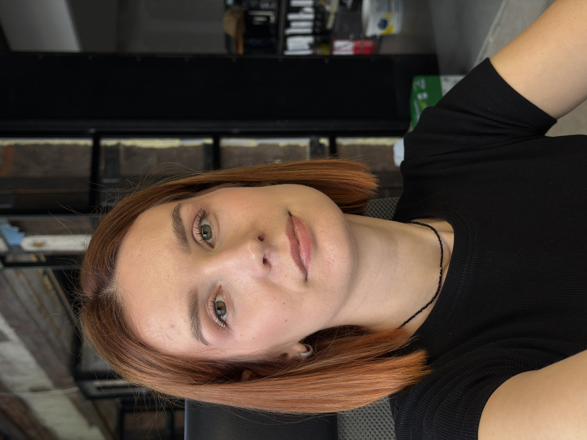 A young woman with auburn, shoulder-length hair is oriented vertically in the frame. She has fair skin, green eyes, and is wearing light makeup. She is wearing a black, short-sleeved shirt, and a delicate, dark necklace is visible around her neck. The background includes an indeterminate interior, potentially a commercial space given the presence of shelving with stacked items. The location is suggested to be Chaikovsky, Russia. The woman is looking directly at the camera.