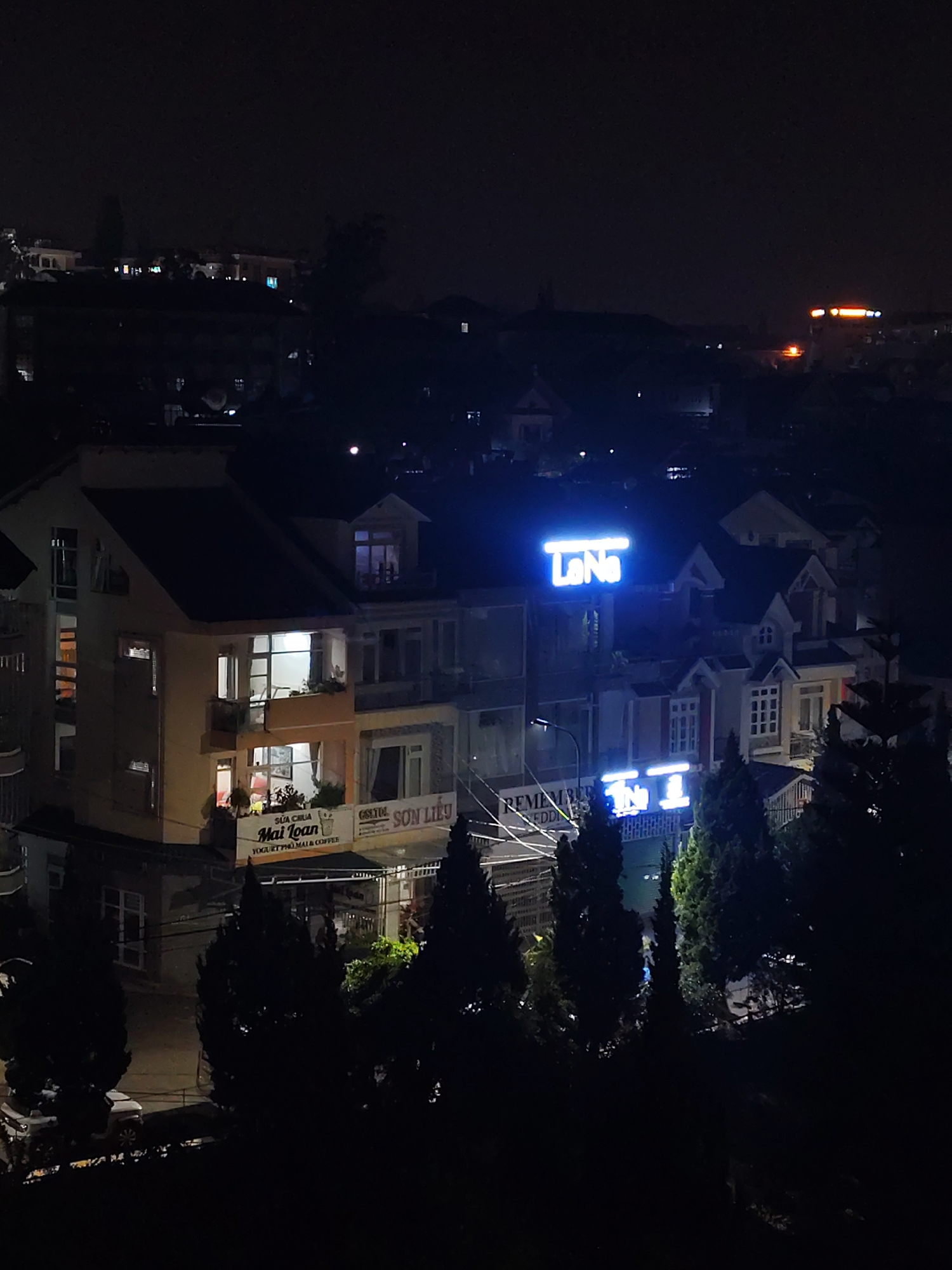The image shows an elevated nighttime view in Da Lat, Vietnam. A building illuminated with a blue neon sign reading "La Na" is a central point, with other structures and residences visible in the background. Street lighting is evident along with a few other illuminated signs, including "Mai Toan Yogurt". Coniferous trees are in the foreground. A parked car is visible on a street level.