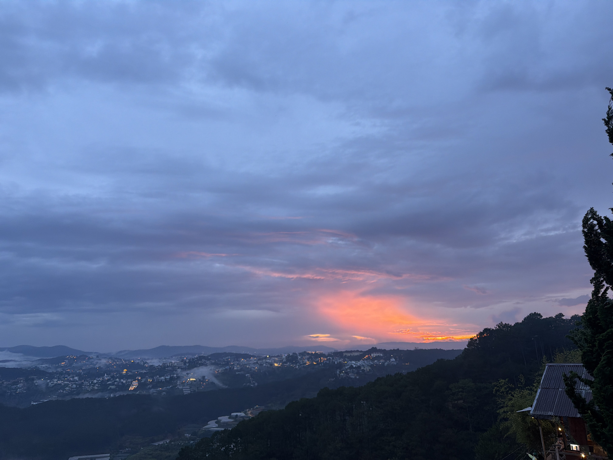 This is a wide-angle, exterior, daytime photograph of a city nestled in a valley at dusk. The sky is dominated by a gradient of blues and grays, with streaks of vibrant orange and pink light originating from the horizon, indicating the setting sun behind the clouds. A soft haze or fog blankets the lower portions of the valley and the distant hills. The city lights are beginning to twinkle, suggesting the transition from day to night. The foreground is characterized by dark, silhouetted trees and a rustic-style building with a corrugated roof on the right side. The city itself appears to be spread across multiple hills, with buildings clustered together, some illuminated by artificial lights. The overall scene captures a serene and picturesque view of Xuan Truong, Da Lat, Vietnam, as twilight descends.