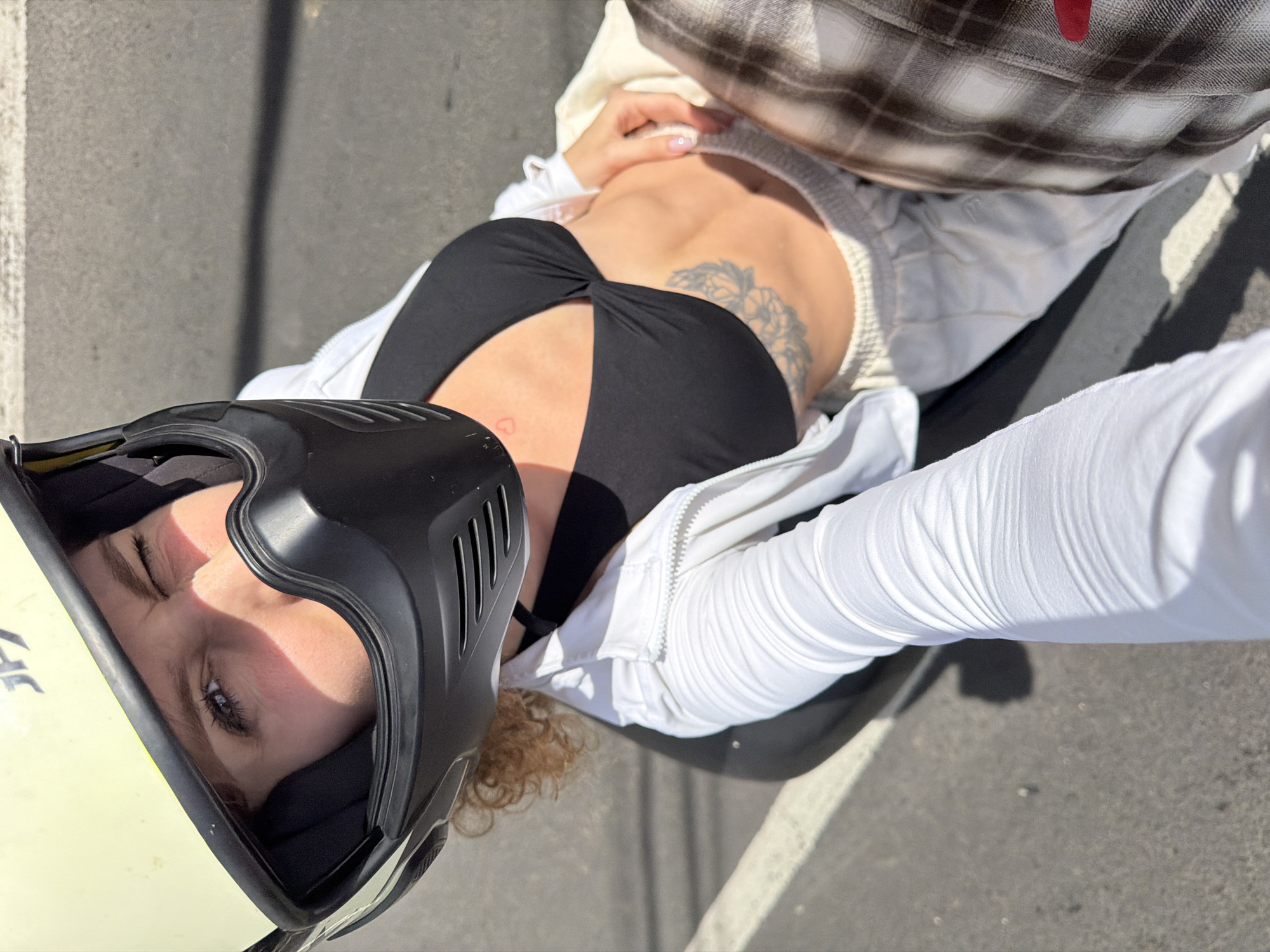 A person wearing a light yellow helmet and a black face mask, a black bikini top, a white zip-up jacket, and light-colored sweatpants is sitting on a motorcycle. The person is winking and looking at the camera. A tattoo is visible on the person's stomach and chest. The person is on a street, indicated by the asphalt and white lines on the ground. The scene is outdoors and illuminated by sunlight, casting shadows.