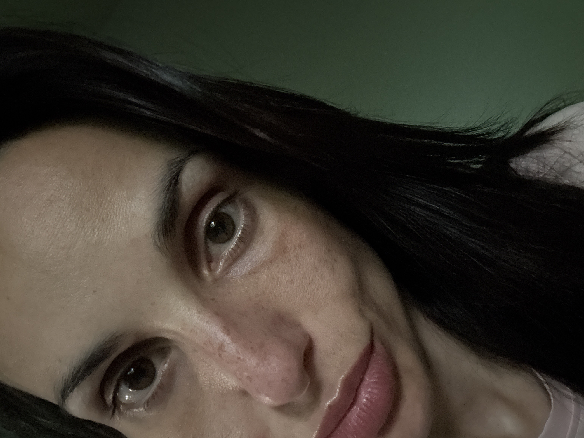 A close-up, slightly angled image shows a portion of a person's face, focusing on the eyes, nose, and mouth. The person has dark hair, fair skin with freckles visible on the nose and cheek, and brown eyes with visible light reflections. The lips are slightly parted and appear to have a gloss or lip color applied. The background is a blurry, dark green. No specific landmarks or details are present that would indicate a location within the city of Pleven, Bulgaria.