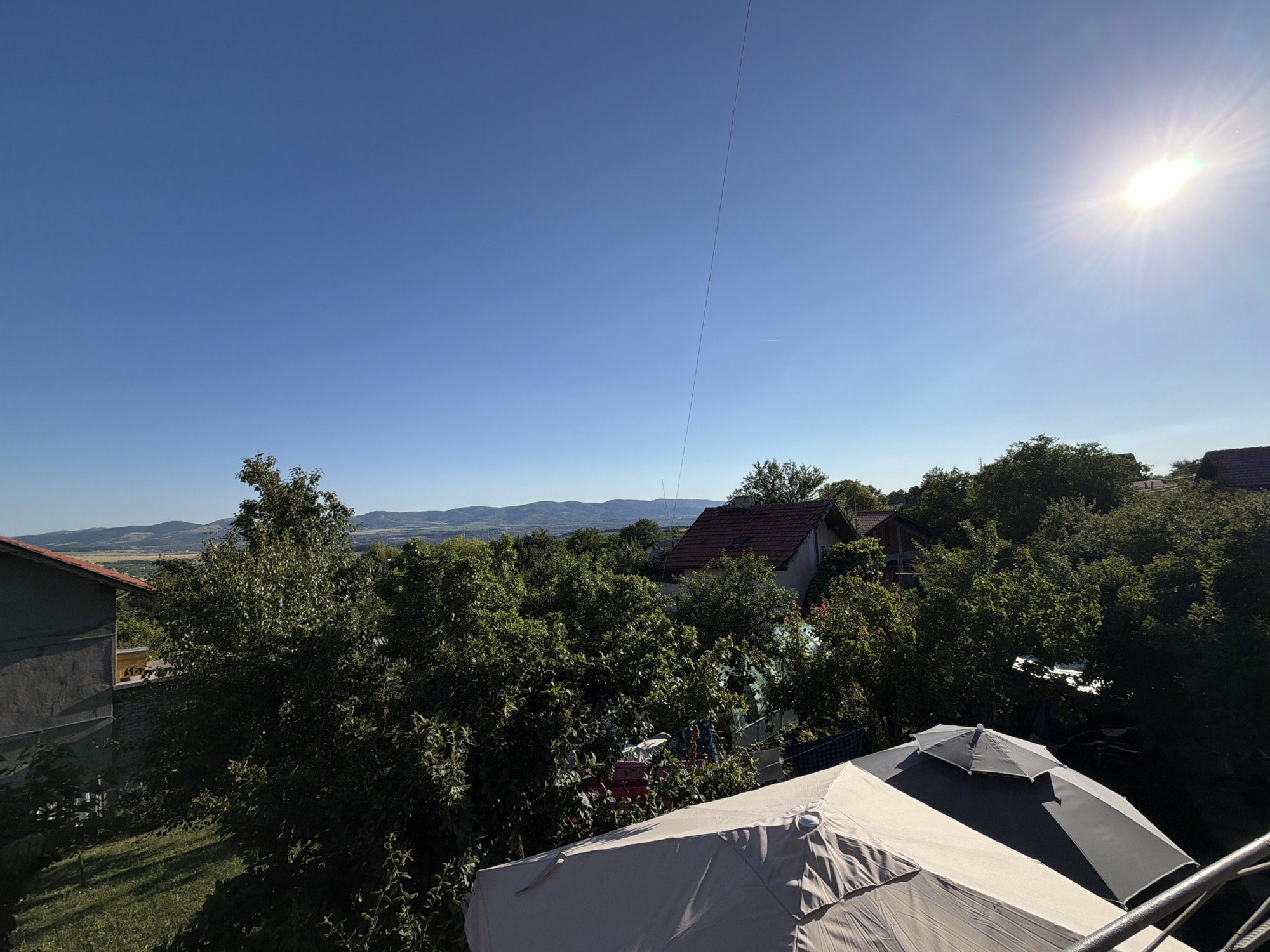 This is a high-angle, wide-shot photograph taken from an elevated position, likely a balcony or a higher floor of a building, overlooking a rural or semi-rural residential area in Golemo Buchino, Bulgaria. The scene is bathed in bright sunlight, with the sun positioned in the upper right corner of the frame, creating a lens flare and casting distinct shadows.
The foreground is dominated by two large outdoor umbrellas. The closest umbrella, in a light beige color, occupies the lower right portion of the image. Partially overlapping it is a darker gray and black umbrella. A metal railing is visible to the far right, suggesting the viewpoint is from a structure with a balcony or deck.
Behind the umbrellas, a dense canopy of green trees fills the midground. Interspersed within the foliage are glimpses of residential buildings with red-tiled roofs. One building on the left, partially visible, has a dark gray, textured facade and a red-tiled roof. Further into the scene, the landscape transitions into rolling hills and mountains in the distance, under a clear, deep blue sky. A thin, dark wire runs diagonally across the sky from the top center towards the right.
The overall impression is one of a sunny day in a village setting, with a focus on outdoor living spaces and the natural surroundings. There are no people or animals clearly visible in the image. The image captures a serene, daytime view of Golemo Buchino.