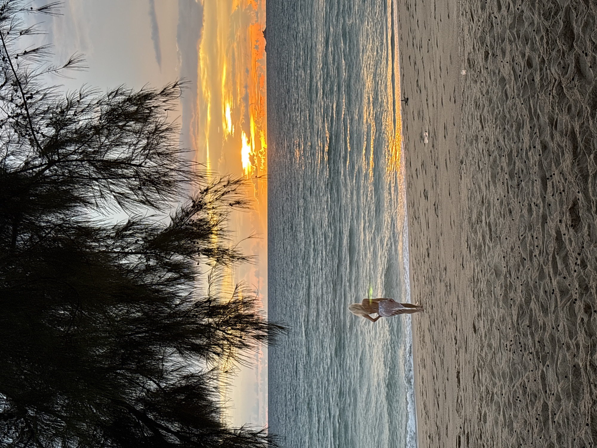 A lone blonde woman stands on a sandy beach facing a calm ocean as a vibrant sunset paints the sky with hues of orange and yellow. She is wearing a patterned wrap skirt over a swimsuit. The water is a deep blue, reflecting the colorful sky. In the foreground, tree branches obscure the top portion of the frame, casting shadows on the beach. The scene appears to be located in Choeng Thale, Thailand, a popular tourist destination known for its beautiful beaches and sunsets.