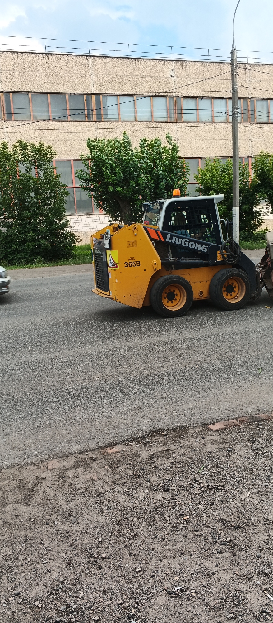 A yellow LiuGong skid steer loader is parked on a paved road in Izhevsk, Russia, next to a large industrial building with rows of windows. The loader has a safety cage and an orange flashing light on top. There is a license plate visible on the front of the loader, reading "365B" and a triangular warning sign with a person and a shovel inside. Trees and bushes are growing between the building and the road. The sky is overcast with some clouds. The road surface is asphalt with some gravel and debris on the side.