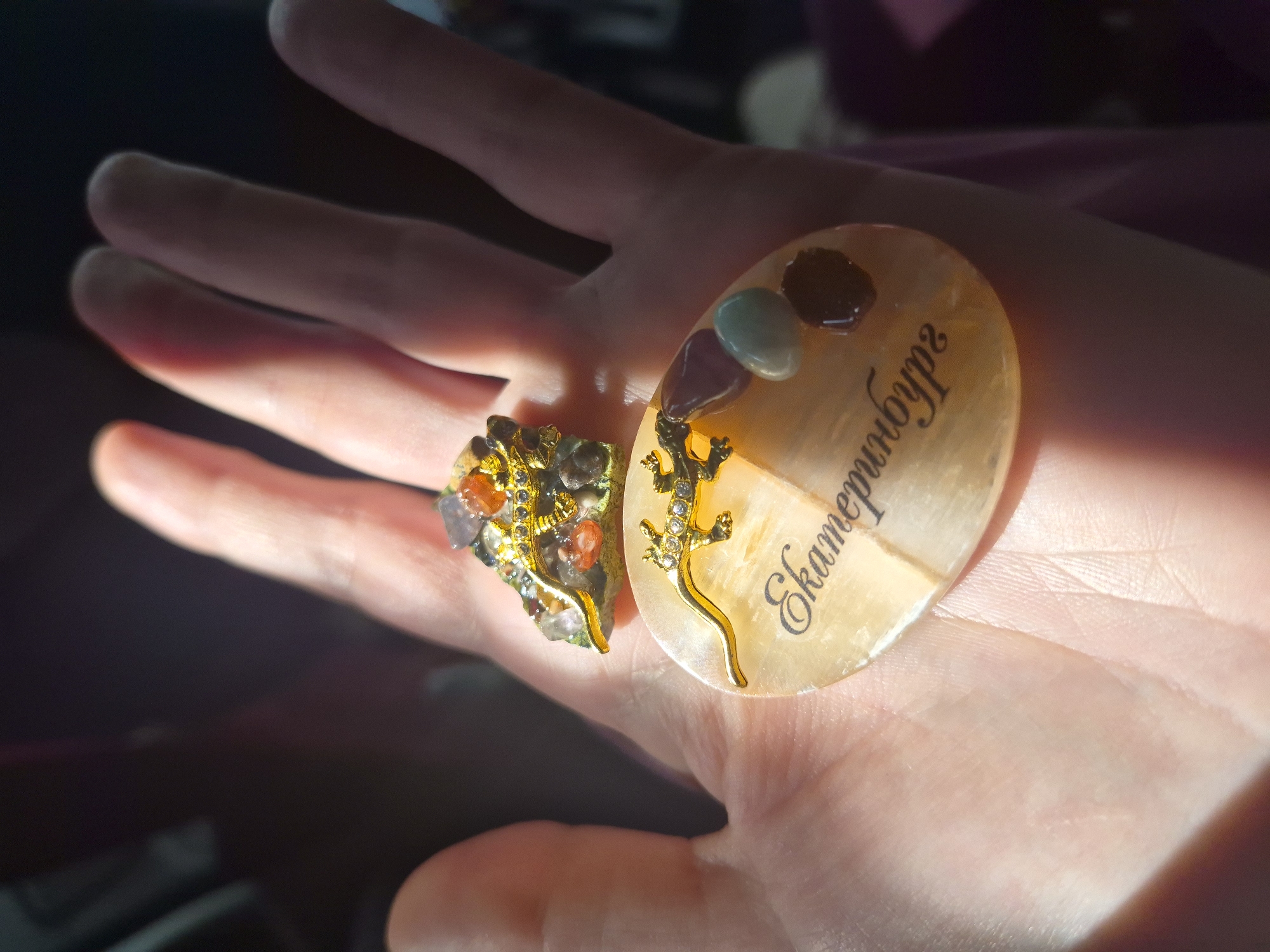 An overhead, close-up shot captures a person's open palm displaying two decorative items. On the palm rests an oval-shaped, translucent light orange stone, which features black cursive text that reads "Екатеринбург" (Yekaterinburg). A golden lizard with small, clear rhinestones adorns the surface of this oval stone. Above and slightly behind the oval stone, a smaller, irregularly shaped cluster of colorful stones, including purple, orange, and clear crystals, is mounted on a green base. This cluster is also adorned with a golden lizard with rhinestone accents. The background is dark and blurred, with shafts of sunlight illuminating the hand and the objects. The image is a close-up view of a hand in Novoalekseevskoe, Russia, holding two decorative stones, one of which is inscribed with the name of the city Yekaterinburg.
