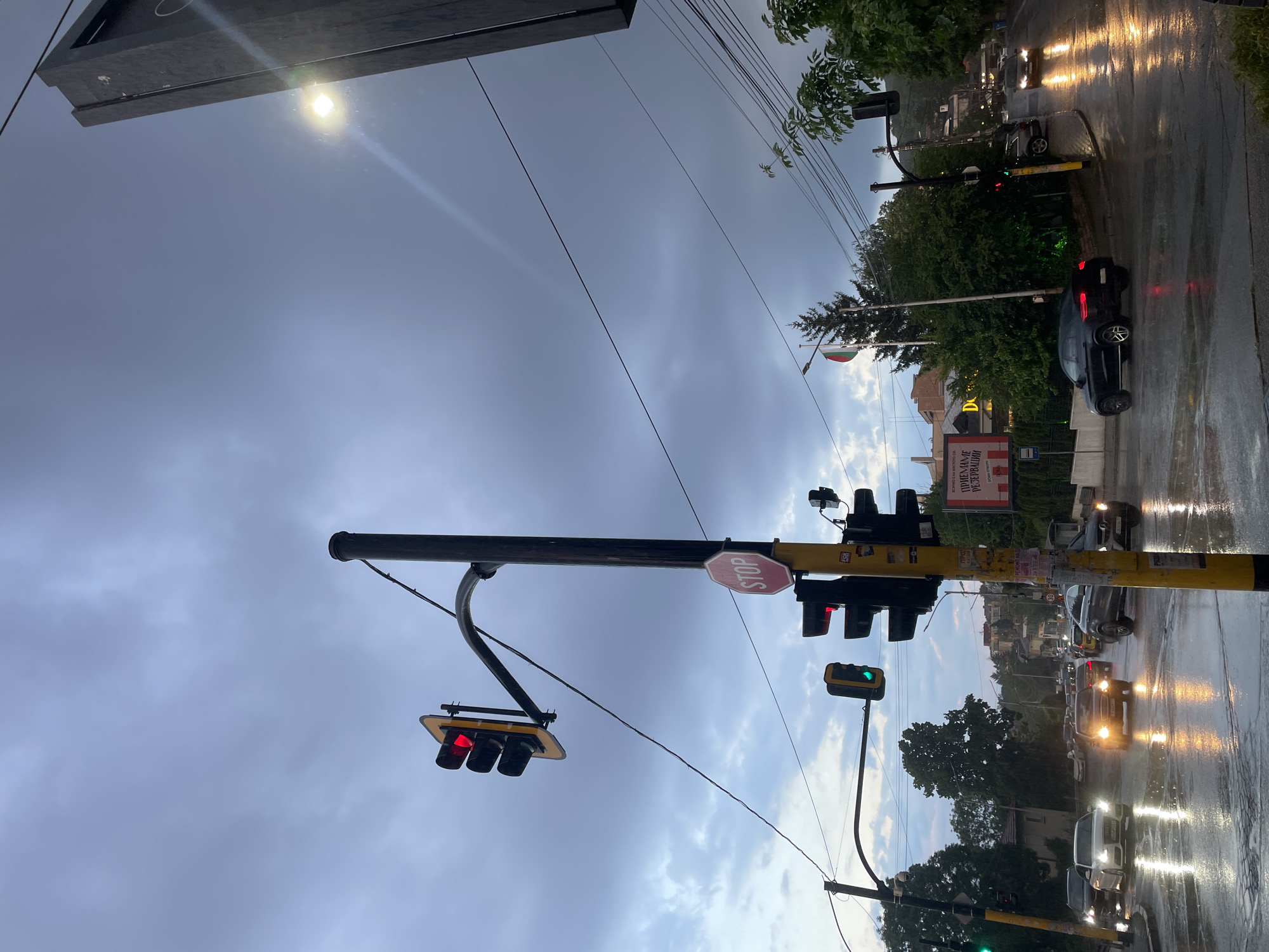 Captured from a low-angle perspective, the image portrays an urban street scene following rainfall, as suggested by the wet road surfaces reflecting the headlights of passing vehicles. Dominating the foreground is a traffic signal pole with red lights illuminated on multiple signals, accompanied by a stop sign. The background reveals buildings and trees amidst a cloudy sky, with the national flag of Bulgaria visible in the distance. The location is Unknown, Unknown.