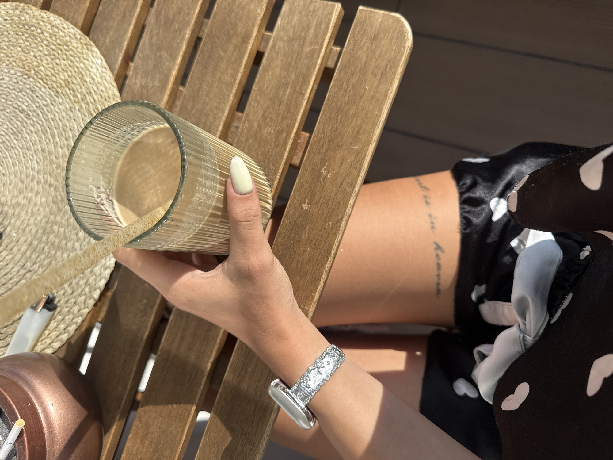 The image shows a person's hand holding a glass of iced coffee on a wooden table. The hand has a light yellow nail polish and is wearing a sparkly watch band. In the background, there is a straw hat and an ashtray with cigarettes. The scene appears to be outdoors, possibly on a patio or balcony, with sunlight casting shadows on the table. The person is wearing a black and white heart-patterned garment. There is a tattoo on the person's leg which reads "God is in heaven". This scene could be located in Plovdiv, Bulgaria.