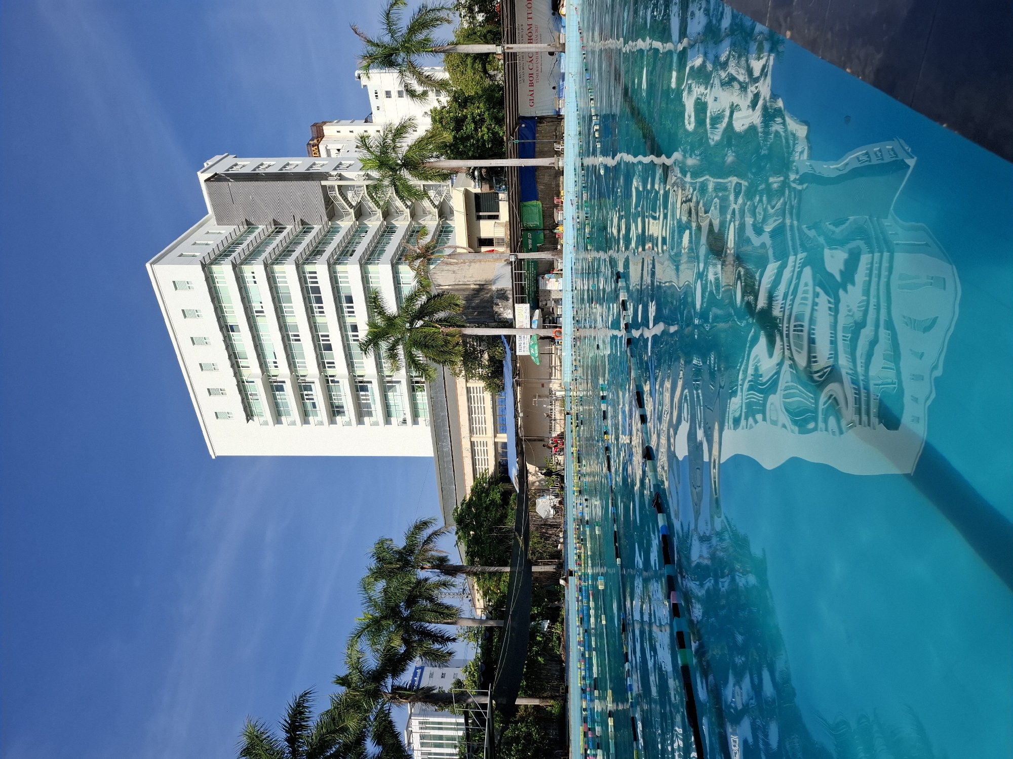 The image depicts an outdoor swimming pool in Nha Trang, Vietnam, under a clear blue sky. The foreground is dominated by the bright blue water of the pool, with lane markers clearly visible. The water reflects the surrounding environment, including a tall white building with multiple windows, palm trees, and the sky. In the background, the white building stands out, with several other smaller buildings and lush greenery visible. There are some people in the background, near the edge of the pool, but they are not the main focus. A banner with Vietnamese text is also visible along the edge of the pool. The overall scene suggests a sunny day at a recreational facility.