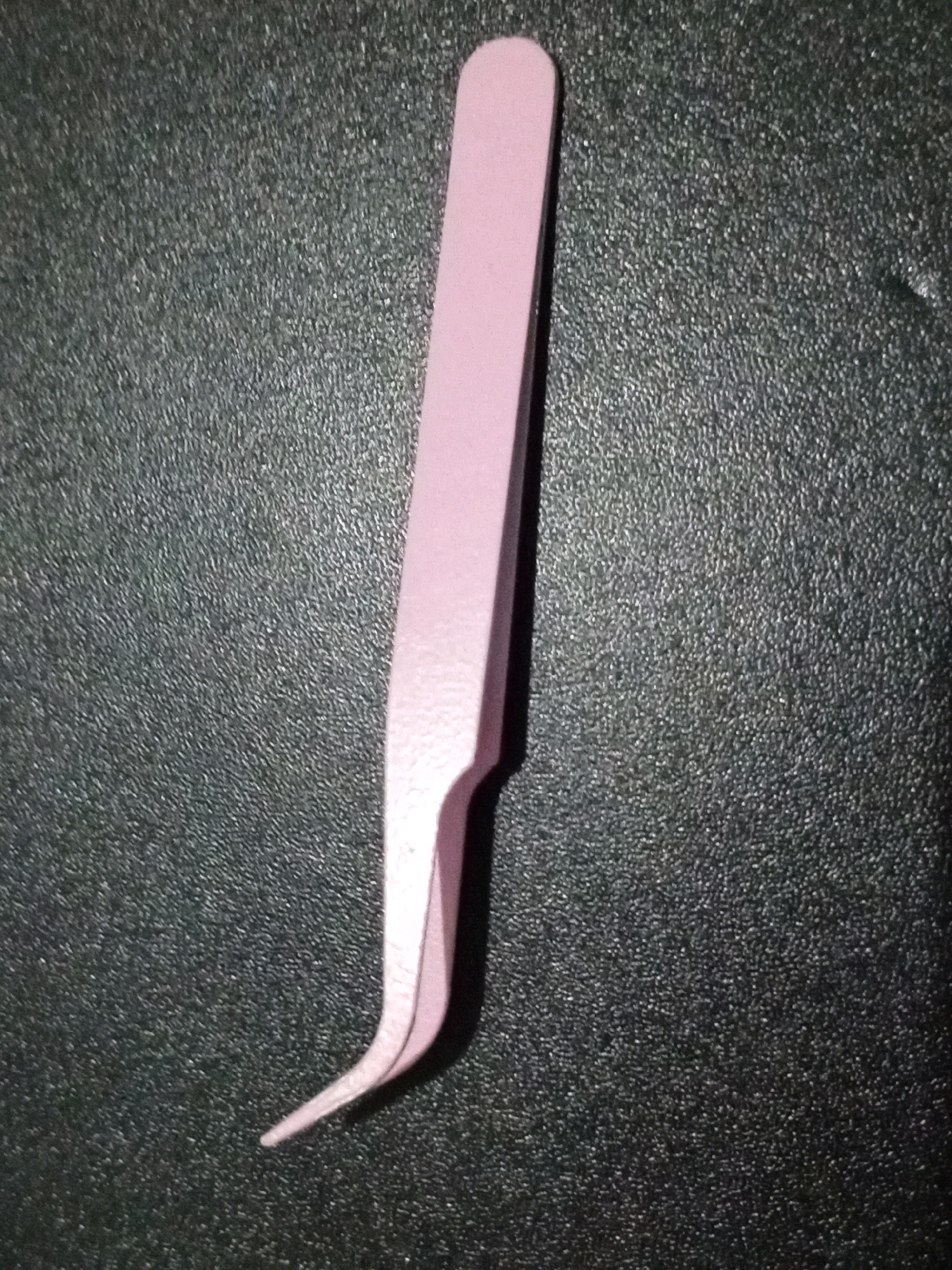 A single, pale pink tweezer lies diagonally on a dark, textured surface. The tweezer's handle is at the top, tapering to a point at the bottom where the tips are curved inward. The background is uniformly dark and appears to be a rough, granular material. There is no indication of the city of Denpasar, Indonesia, or any other location. The image is a close-up shot of the object.