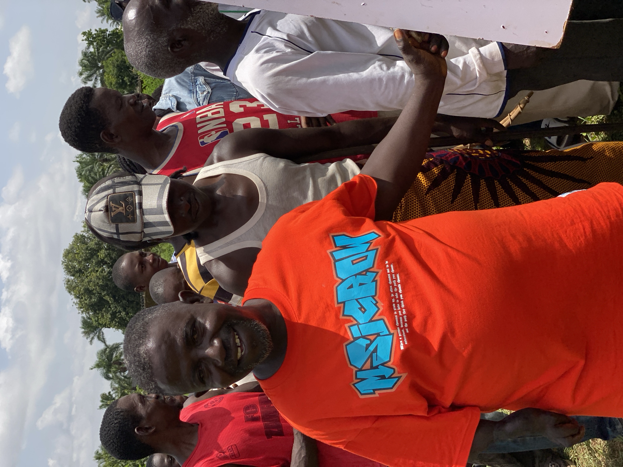 In Maigizo, Nigeria, a group of men stand closely together outdoors under a partly cloudy sky. In the foreground, a man in an orange t-shirt with the word "MSICACK" printed in blue leans towards the camera, smiling. To his left, a man wears a red jersey. Behind them, individuals wear a white tank top, a cap with a logo, and an NBA jersey, among other clothing. Some are holding onto a white, rectangular object, possibly a sign or board. Trees are visible in the background, suggesting a natural environment within the city.