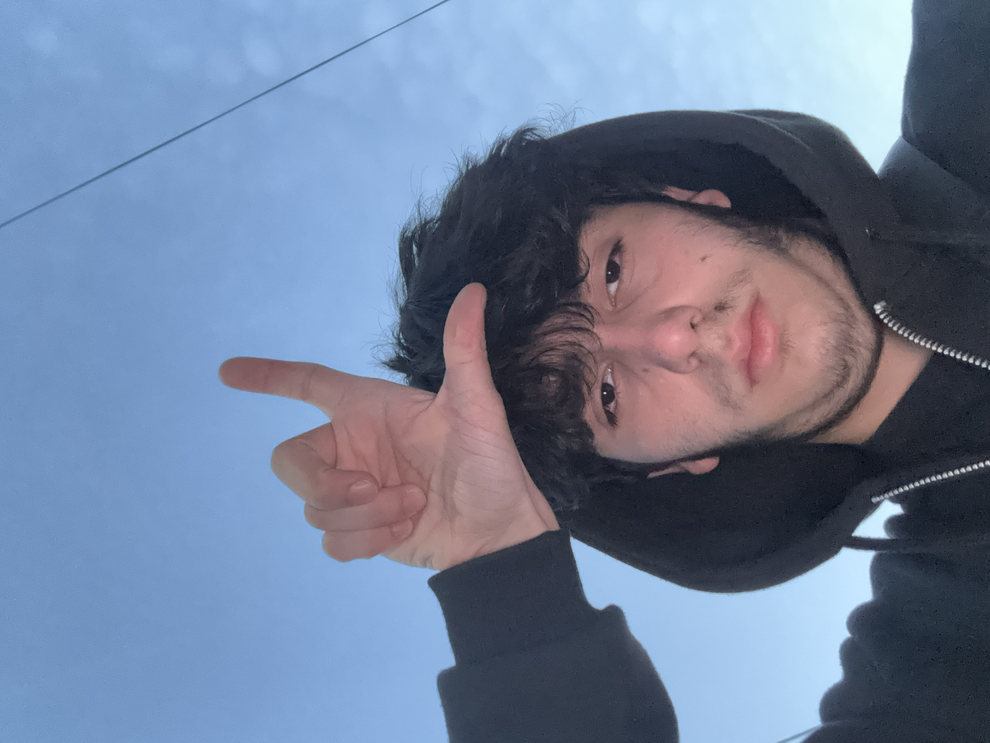 A selfie from a low angle shows a young man wearing a black hooded sweatshirt against a pale blue sky. He is making a hand gesture, with his index finger extended and thumb raised, pointing towards the upper left of the frame. The man has dark, wavy hair that falls partially across his forehead. He has light skin, dark eyes, and a light beard. The photo was taken outdoors, as indicated by the clear sky. No buildings or landmarks are visible. There is no indication of the location of this picture as being in Yerevan, Armenia.