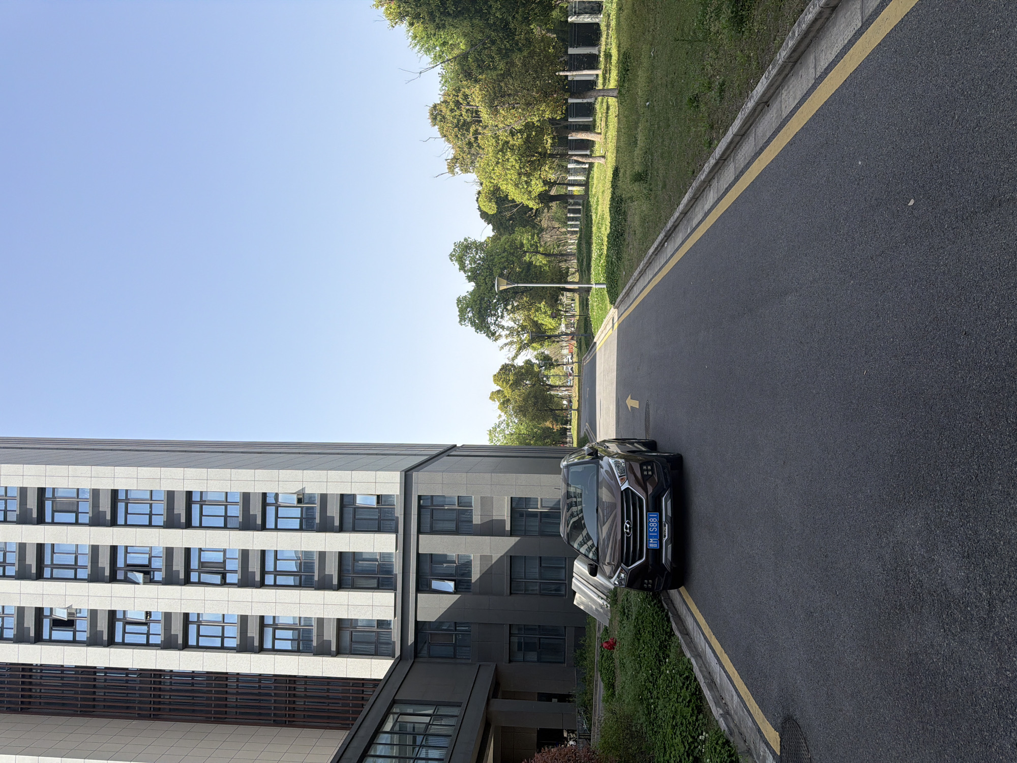 The image shows a black Hyundai sedan parked on the side of a road next to a modern, multi-story building in what appears to be the Jiangning District of China. The building has a gray and white facade with numerous windows. A yellow line marks the edge of the road, and green grass and trees are visible on the other side of the road. The sky is clear and blue. The license plate on the car is visible.