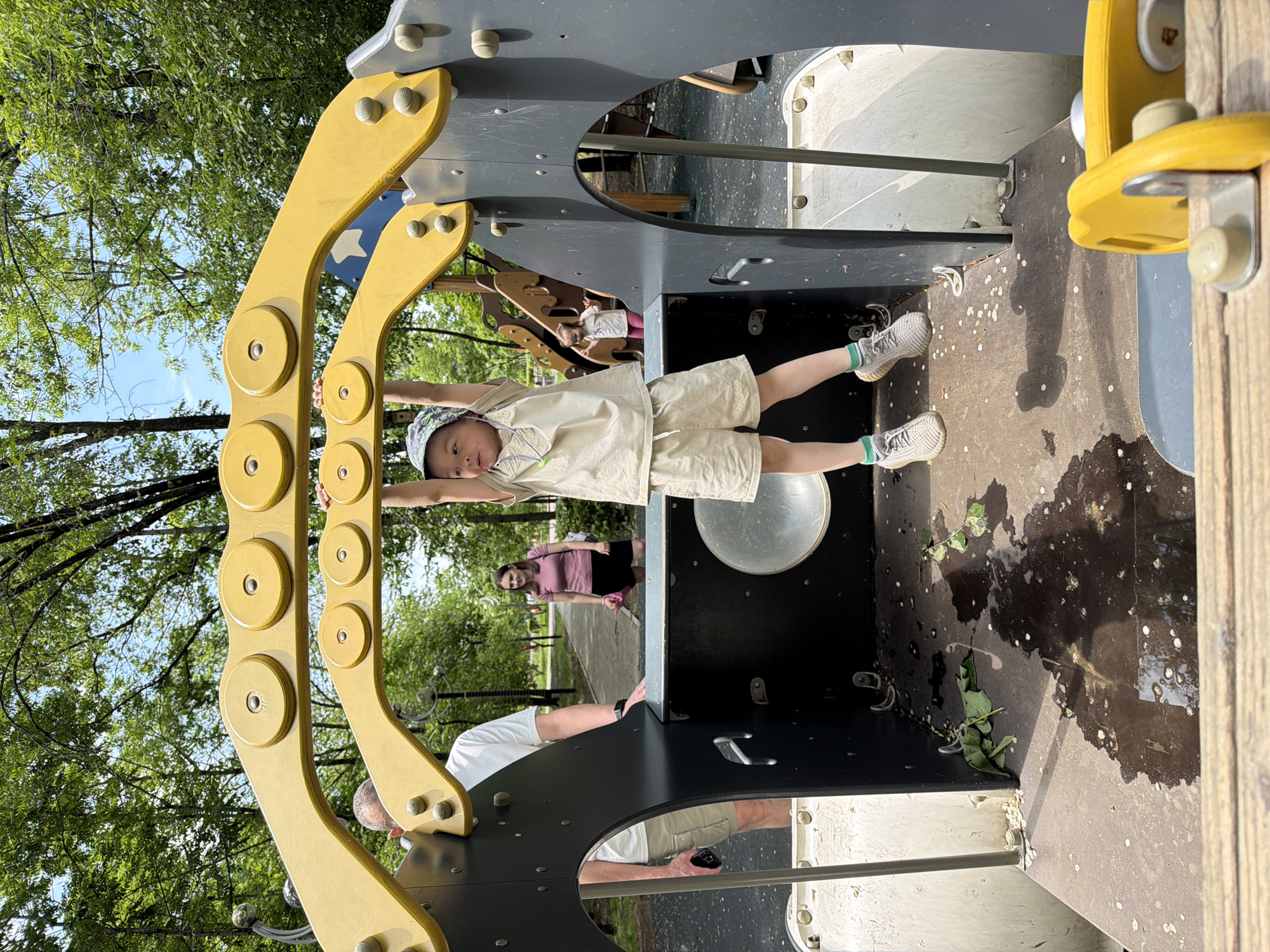 An outdoor shot captures a child on a playground in Khabarovsk, Russia. The child, a boy wearing a light-colored t-shirt, shorts, a patterned cap, and sneakers, is hanging from a yellow overhead structure. He is positioned in the foreground, with his arms extended upwards. Behind him, a man in a white t-shirt and beige shorts stands to the left, and a woman in a pink t-shirt and black shorts is visible further back on a pathway. Another child, a girl in a light-colored t-shirt and pink pants, is on a slide structure in the background. The playground equipment is predominantly gray and yellow, with some wooden elements. The ground appears to be a dark, textured surface, with some wet patches and scattered leaves. Trees with green foliage form the backdrop against a clear blue sky.