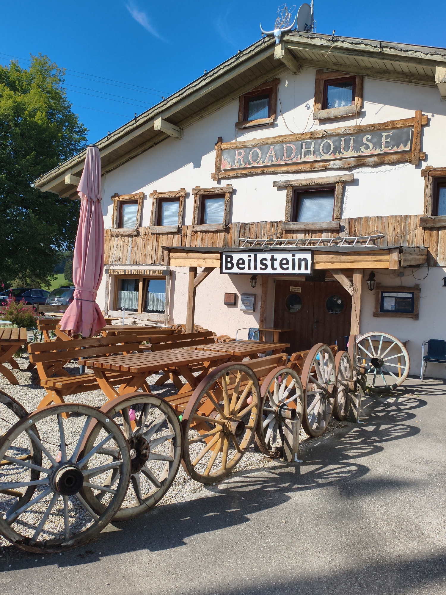 This is a full shot of a building with a sign that says "ROADHOUSE" and another sign that says "Beilstein". There are several wooden picnic tables and benches in front of the building, and a pink umbrella is folded up next to one of the tables. In the foreground, there are several large, old-fashioned wagon wheels. The building appears to be a restaurant or bar, possibly in a rural setting. The sky is blue with a few wispy clouds. The building is made of wood and stucco and has several windows. The overall impression is one of a rustic, casual establishment. The location is Stötten am Auerberg, Germany.