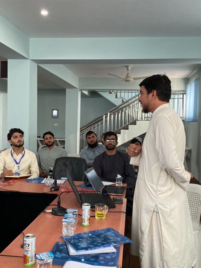 **Location:** Shurandam, Afghanistan
**Scene:** An indoor meeting or presentation is in progress. Five men are seated at a reddish-brown table covered with papers, cans of juice, and water bottles. Two open laptops are on the table. The men at the table are facing a standing man who appears to be addressing them. The seated men are wearing various types of shirts, with one wearing a lanyard with an ID badge. The standing man is wearing a long, light-colored garment. Behind them is a stairwell. The room has light blue walls. There is a ceiling fan.