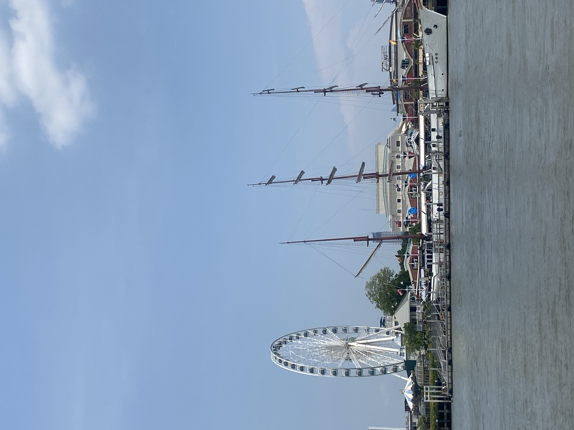 The image captures a scene in Bangkok, Thailand, showcasing the Chao Phraya River waterfront. Dominating the view is a large Ferris wheel, likely the "Asiatique The Riverfront" Ferris wheel, a notable landmark along the river. Several tall-masted ships are docked along the waterfront, suggesting a tourist or leisure activity. A building or structure, possibly part of the waterfront development, is visible behind the ships. The sky is a clear blue with some scattered clouds. The foreground shows the edge of a pier or riverbank.