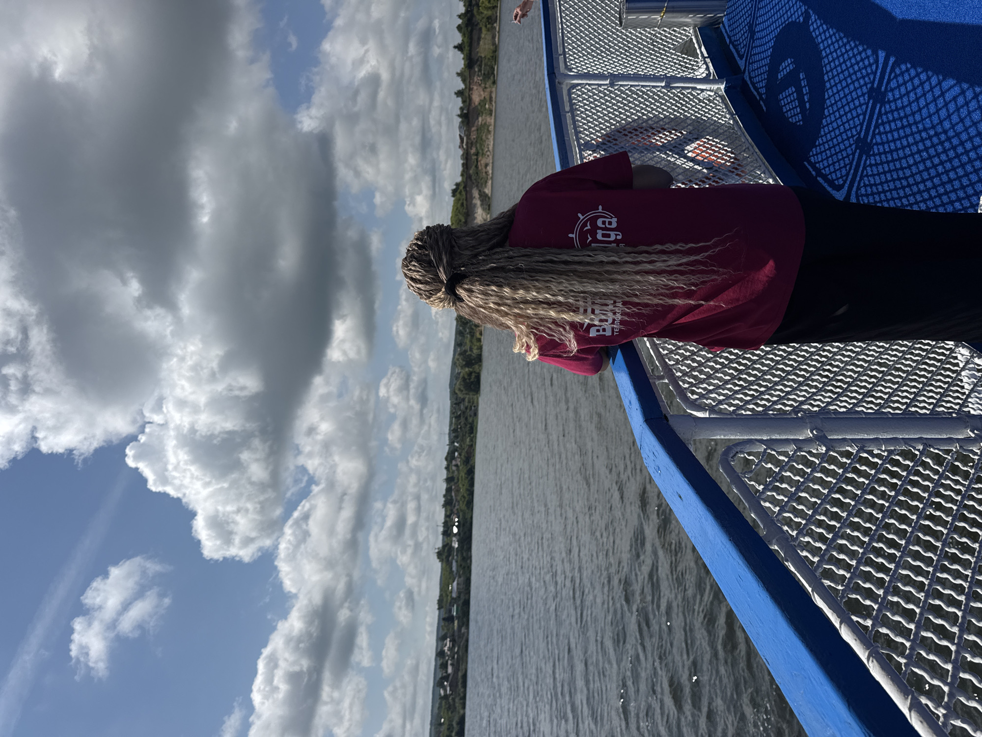 The image depicts a woman with blonde braided hair standing on the deck of a boat, looking out at a wide river. The boat has a blue railing and a white metal fence. The river is calm and reflects the sky. In the distance, across the river, a village is visible on a treed embankment. The sky is partly cloudy with dramatic, large white and gray clouds against a blue sky. The woman is wearing a dark red t-shirt with a circular logo on the back, and a long black skirt. The location is the city of Shelanga, Russia.