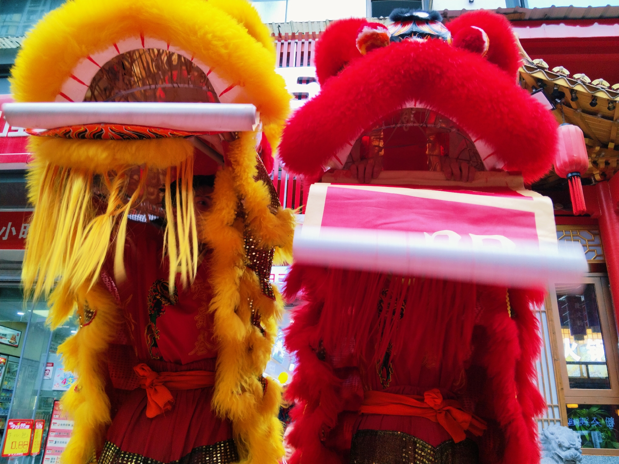 Two lion dance costumes, one yellow and one red, are the central subjects, obscuring the faces of the performers within. The yellow costume is on the left and the red on the right. Both are ornately decorated with vibrant fur and traditional patterns. The costumes feature waist sashes and headpieces with semicircular frames.
The background shows an urban environment, likely in the Yuzhong District of China. Visible elements include storefronts, a building with red accents and a red lantern, and glimpses of signs with Chinese characters. The lighting suggests daytime.