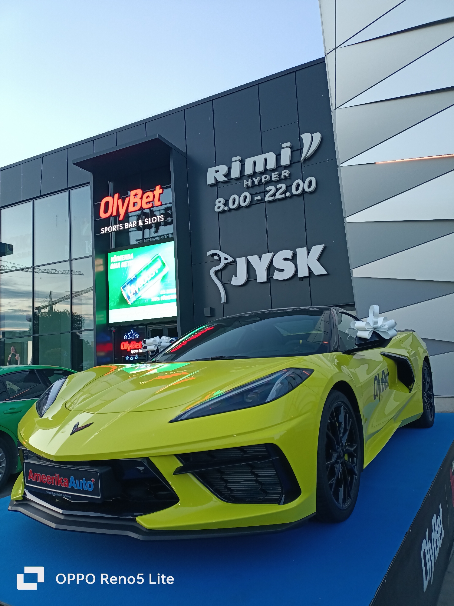 The image shows a bright yellow Chevrolet Corvette C8 parked on a blue platform in front of a modern building. The building has large glass windows and a dark gray facade with illuminated signs. One sign reads "OlyBet SPORTS BAR & SLOTS," and another, higher up, displays "Rimi HYPER 8.00-22.00." Below the Rimi sign is a white "JYSK" logo. A large green can advertisement is displayed on a screen in the window. To the left of the Corvette, a green car is partially visible. The Corvette has a large white bow on its windshield, suggesting it might be a prize or gift. The license plate area of the Corvette displays "AmeerikaAuto" with a website address. The scene appears to be an outdoor promotional event or display in a commercial area. The overall lighting suggests it is daytime, likely late afternoon or early evening. The building's architecture, particularly the angular panels on the right side, suggests a contemporary design. The presence of cranes in the background, visible through the glass, indicates ongoing construction in the vicinity.