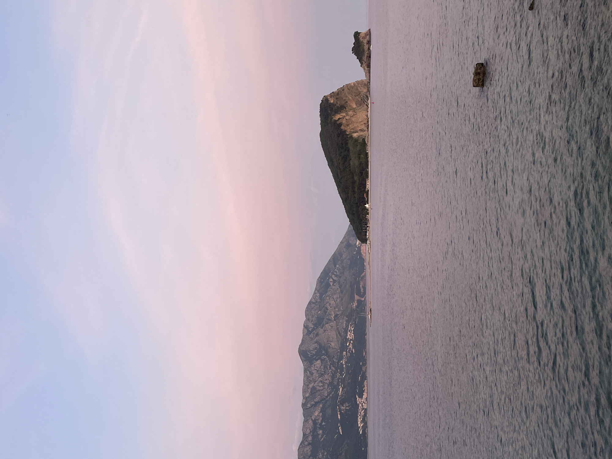 The image depicts a coastal scene in Unknown, Unknown. On the left, the sky transitions from blue to pink. Adjacent to the sky is a mountain range with a town visible along its base. To the right of the mountains is the ocean. In the distance, a small rocky island is present. There's a small floating object in the ocean.