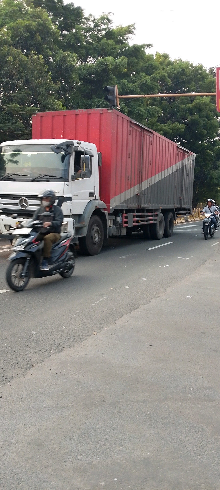 This is a street-level, eye-level, vertical, medium shot captured in Depok, Indonesia, depicting traffic on a paved road. The scene is dominated by a large, white and red semi-trailer truck with a red container, positioned on the right side of the road and moving away from the viewer. To the left of the truck, a person wearing a helmet and mask rides a black scooter towards the viewer. Further to the right, another person on a motorcycle, also wearing a helmet, is seen moving away from the viewer. The road surface is asphalt, with visible white lane markings. In the background, lush green trees create a dense canopy, and a traffic light is visible on a pole above the truck. The sky is overcast. The image appears to have been taken during daylight hours.