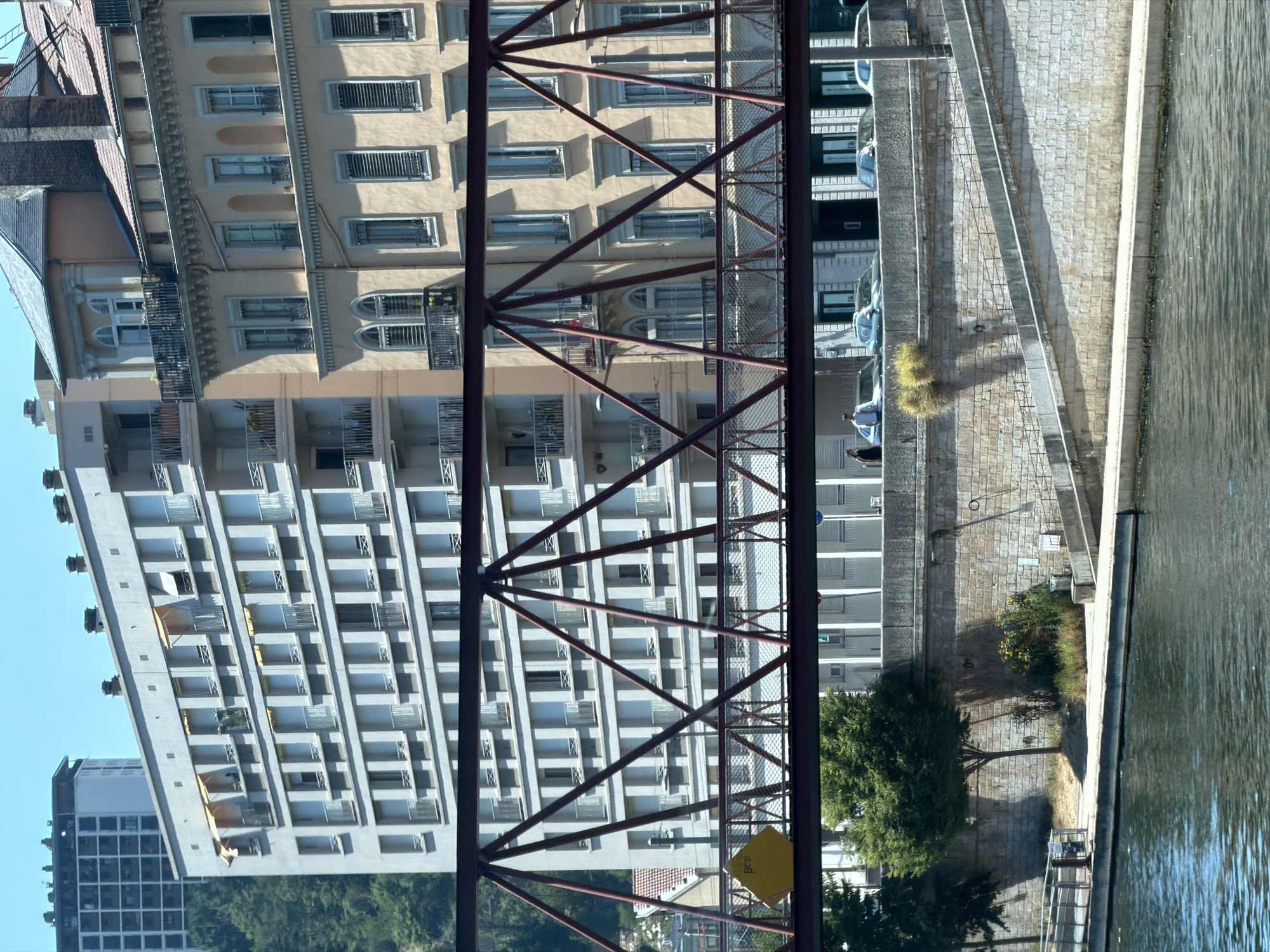 The image shows a view of the Rhône River in Lyon, France, with a bridge in the foreground and buildings on the opposite bank. The bridge is a metal truss bridge painted a dark reddish-brown. A pedestrian walkway with a yellow hexagonal sign is visible on the bridge. Behind the bridge, there are two large buildings. The building on the left is a modern, light-colored apartment building with many windows and balconies. To the right of that is an older, cream-colored building with ornate architectural details, including arched windows and decorative cornices. The river is wide and appears calm, reflecting the blue sky and the buildings. A stone embankment with a paved walkway lines the river. In the middle ground, on the walkway, two people are visible, one in a light blue shirt and the other in a dark top. Several cars are parked along the street behind the walkway. Lush green trees are visible on the left side of the image, obscuring some of the lower parts of the buildings and embankment. The overall scene suggests a sunny day in an urban environment.
