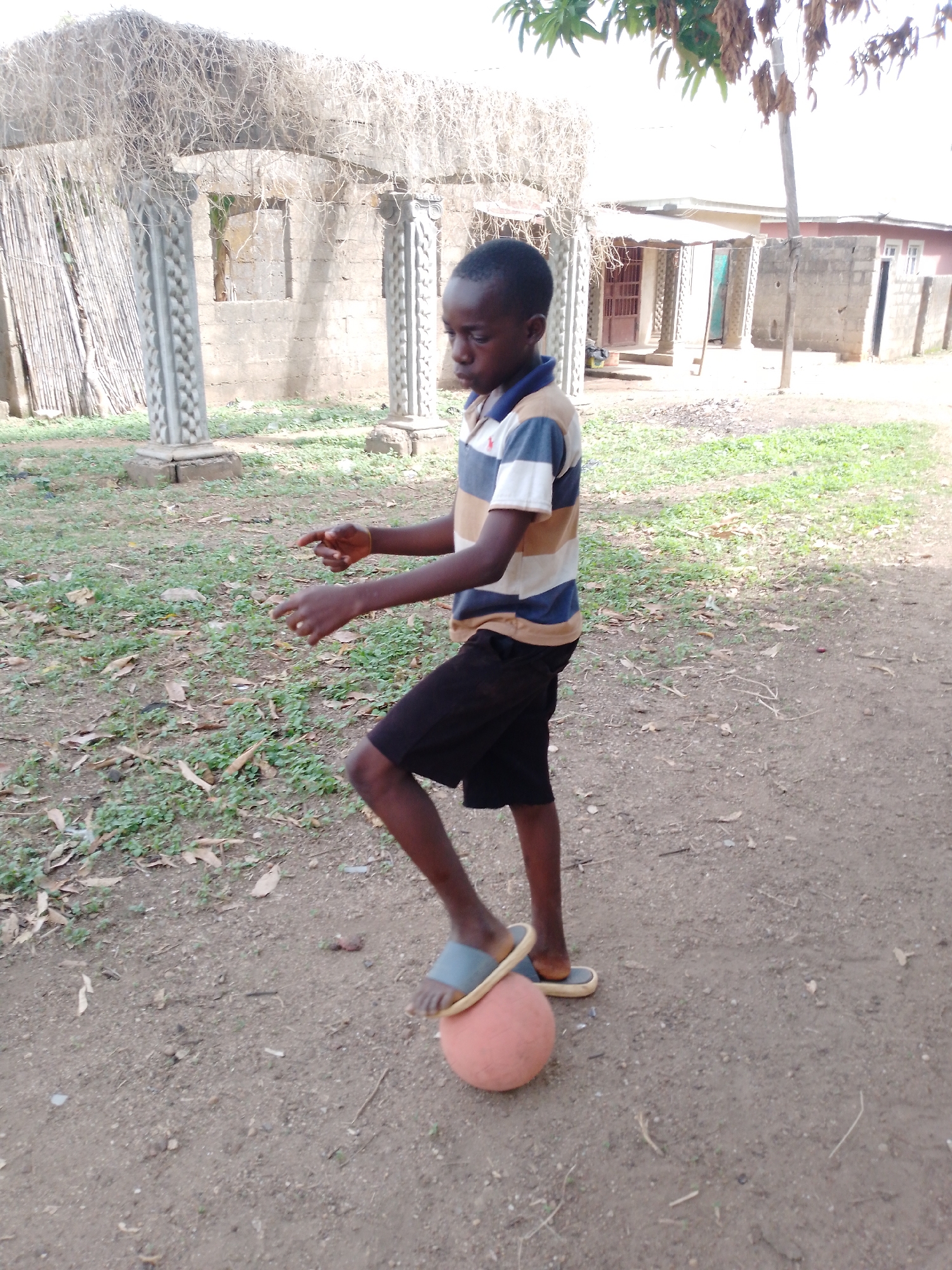 A young boy, likely of African descent, is captured in an outdoor setting, balancing an orange or reddish-orange ball with his left foot. He wears a horizontally striped polo shirt in blue, white, and tan, featuring a small red logo on the left chest, paired with black shorts and light blue flip-flops. His hands are slightly raised and open, suggesting concentration or a moment in play. The boy's gaze is directed downwards at the ball.
The scene unfolds on an unpaved dirt ground, sparsely covered with green patches of grass and scattered dry leaves. In the background, to the boy's left, stands a structure featuring two decorative concrete pillars with intricate carvings, topped by a canopy of dry, stringy vegetation, possibly vines or straw. Behind this, an unfinished concrete block building with open window frames and a wooden fence or stack of bamboo is visible. Further back, another house with a reddish roof and light-colored walls can be seen, along with a vertical wooden pole. The bright, natural light indicates it is daytime. The setting appears to be a rural or semi-urban area. No legible text is visible in the image.