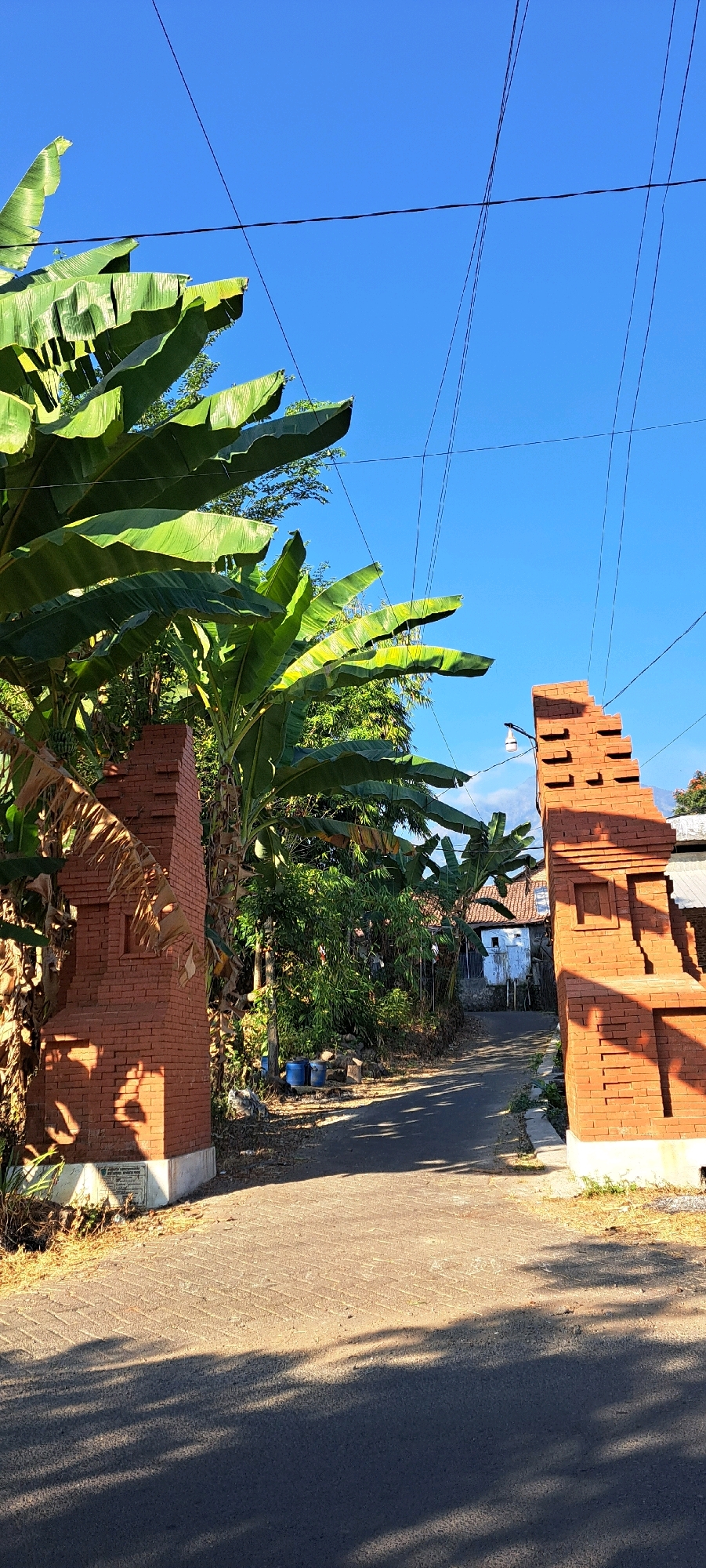 The image shows a sunlit, narrow asphalt road in Sukolelo, Indonesia, flanked by brick gateposts and lush banana trees. The gateposts, constructed from reddish-brown bricks with decorative notches, stand on either side of the road, framing a view of a small house with a white facade and a tiled roof in the distance. Several power lines crisscross the bright blue sky overhead. The ground is a mix of asphalt and textured pavement, with shadows cast by the banana leaves and the gateposts. There are no people or animals visible in the scene. The environment appears to be a rural or semi-rural area.