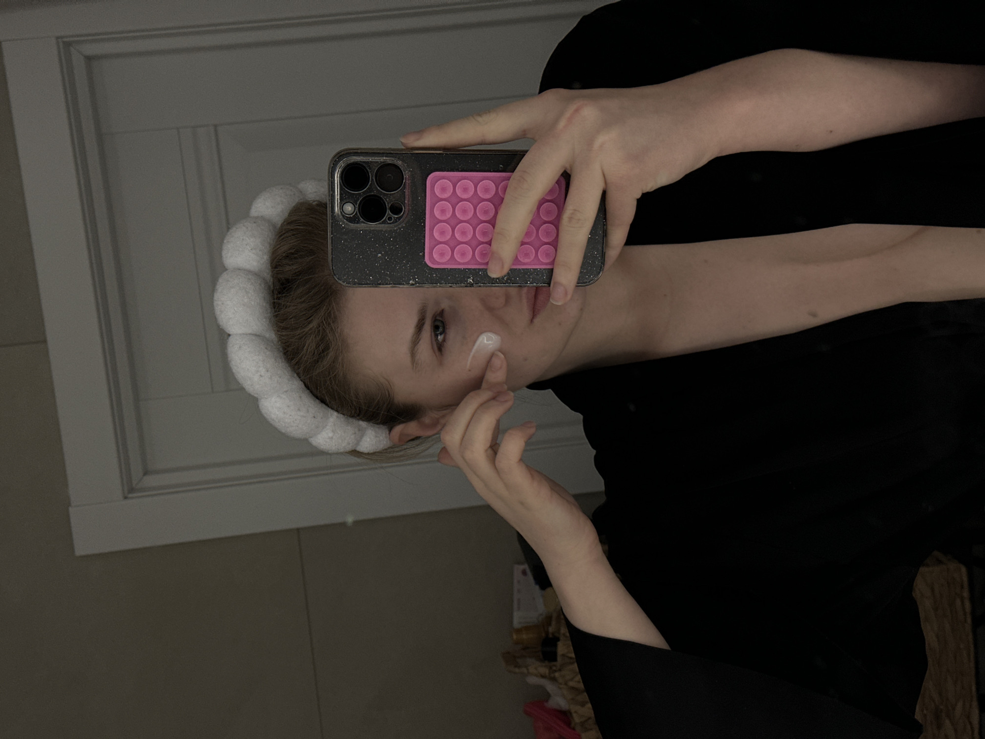 The image depicts a mirror selfie of a young adult female inside an indoor residential setting, potentially located in Kazan, Russia. The subject has light hair and is wearing a white, segmented, puffy headband, with a dark, open robe or garment. She holds a dark-cased iPhone with a pink 'pop-it' style grip in her right hand, capturing the reflection. Her left hand is touching her left cheek, where a small amount of white cream, likely a skincare product, is visible. The background reveals a white paneled door on the upper right, a plain light-colored wall, and a portion of a woven basket containing various items on the lower left.