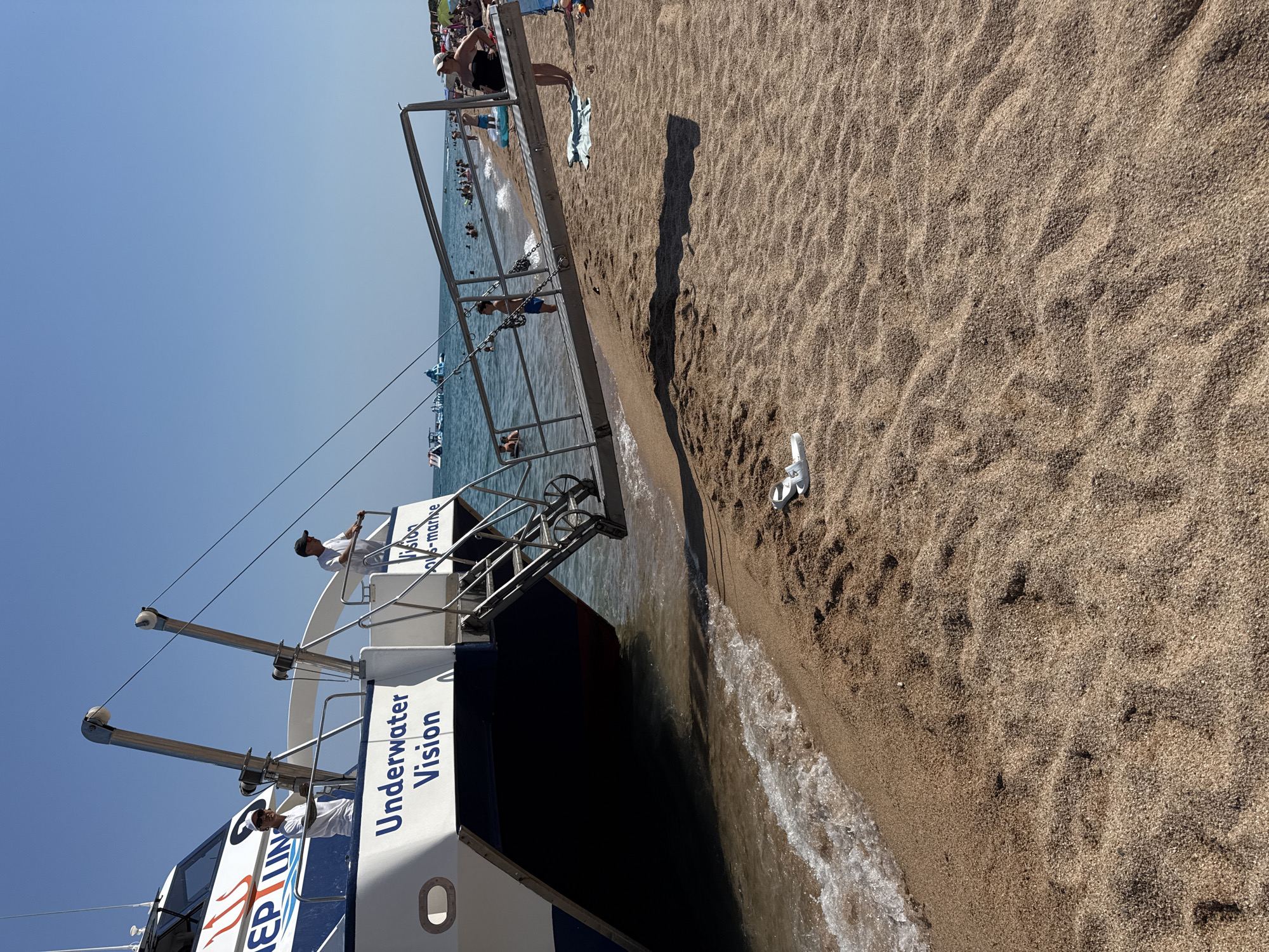 The image depicts a sunny day at the beach in Blanes, Spain, likely near the Blanes Yacht Club, judging by the presence of a large boat. The boat, named "Underwater Vision," is partially submerged at the water's edge, with a ramp extending onto the sandy beach. Two individuals, appearing to be crew members wearing white shirts, stand on the deck of the boat. On the beach, a few people are visible, some swimming or playing in the water, while others are relaxing on the shore. The ocean is calm, with gentle waves lapping at the beach. The sand is light brown and appears somewhat rippled. In the background, a few inflatable water structures are present in the sea, and further down the coastline, beach umbrellas and more people can be seen. The sky is a clear, bright blue, indicating fair weather.