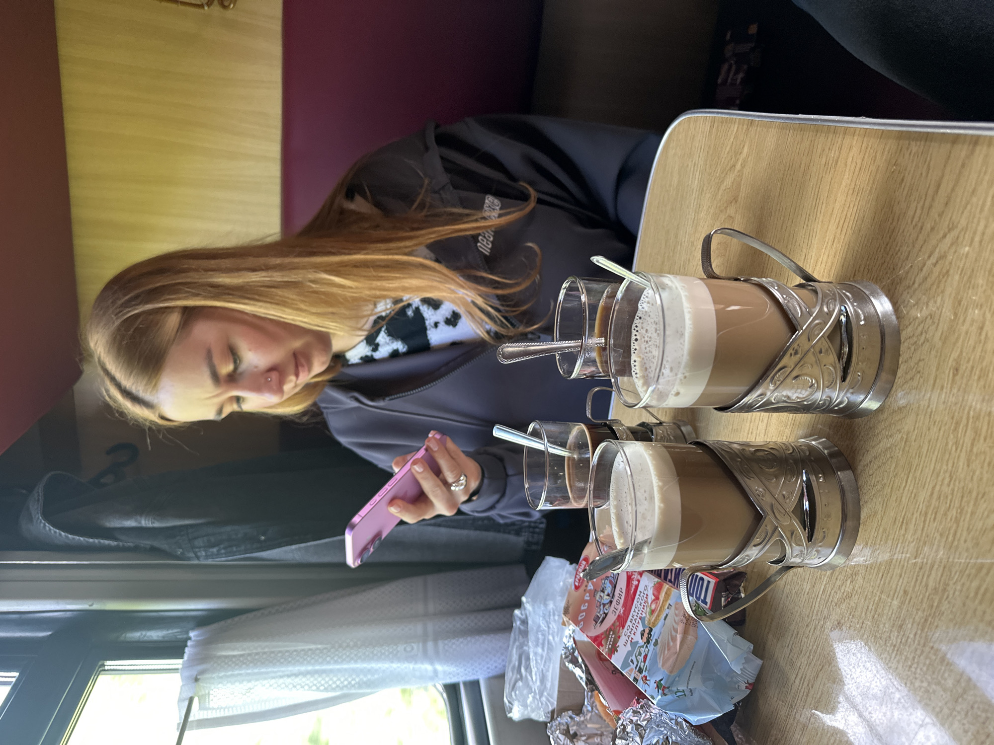 The image shows a young woman with long, light brown hair, wearing a dark jacket over a black and white patterned shirt, sitting on a train. She is looking at a pink smartphone she is holding. On the table in front of her are two glasses of layered coffee or latte in ornate silver holders. There are spoons in each glass and a package of what appears to be pastries or snacks. The train cabin has a wooden table, a window with a white curtain, and seating with dark red upholstery. The location is likely Мажэйкаўскі сельскі Савет, Belarus, given the location context provided.