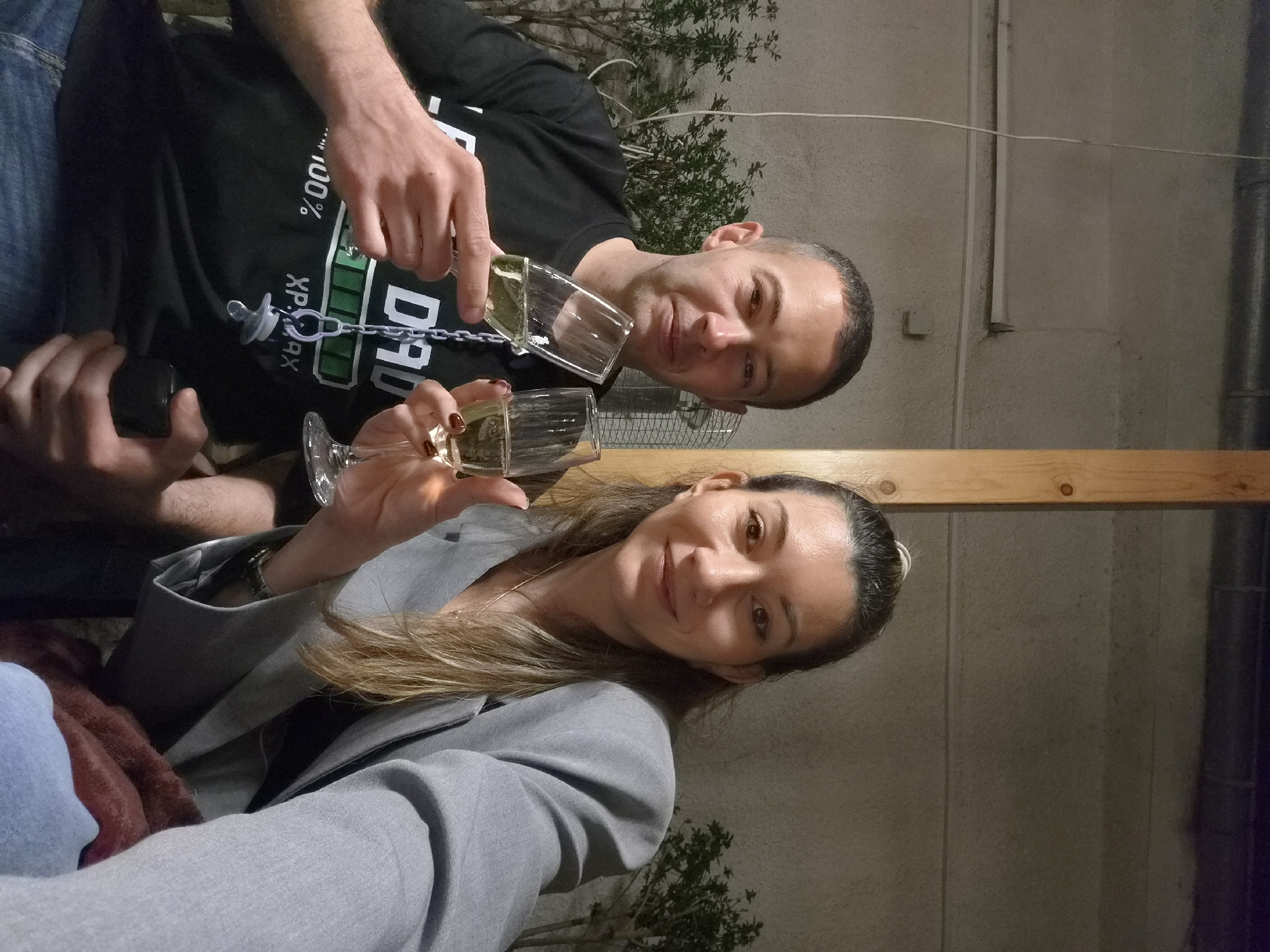 A couple is posing for a selfie, holding champagne glasses. The man wears a black t-shirt with a "DAD 100% XPAX" graphic and a pacifier chain. The woman wears a gray blazer. The location appears to be outdoors, possibly in Plovdiv, Bulgaria, given the local context request, but there are not enough distinct details to confirm the exact location. A wooden beam is visible overhead, and a concrete-like wall is in the background. There is some greenery behind the man. The couple seems to be toasting.