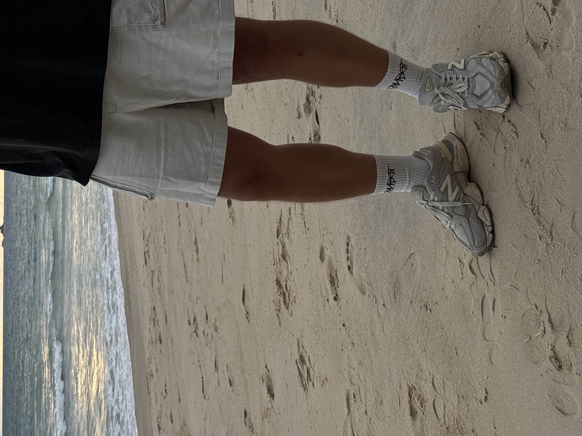 A person stands on a sandy beach in Choeng Thale, Thailand, with the ocean and waves in the background. They are wearing light-colored shorts, white socks with black text, and grey athletic shoes. Footprints are visible in the sand.