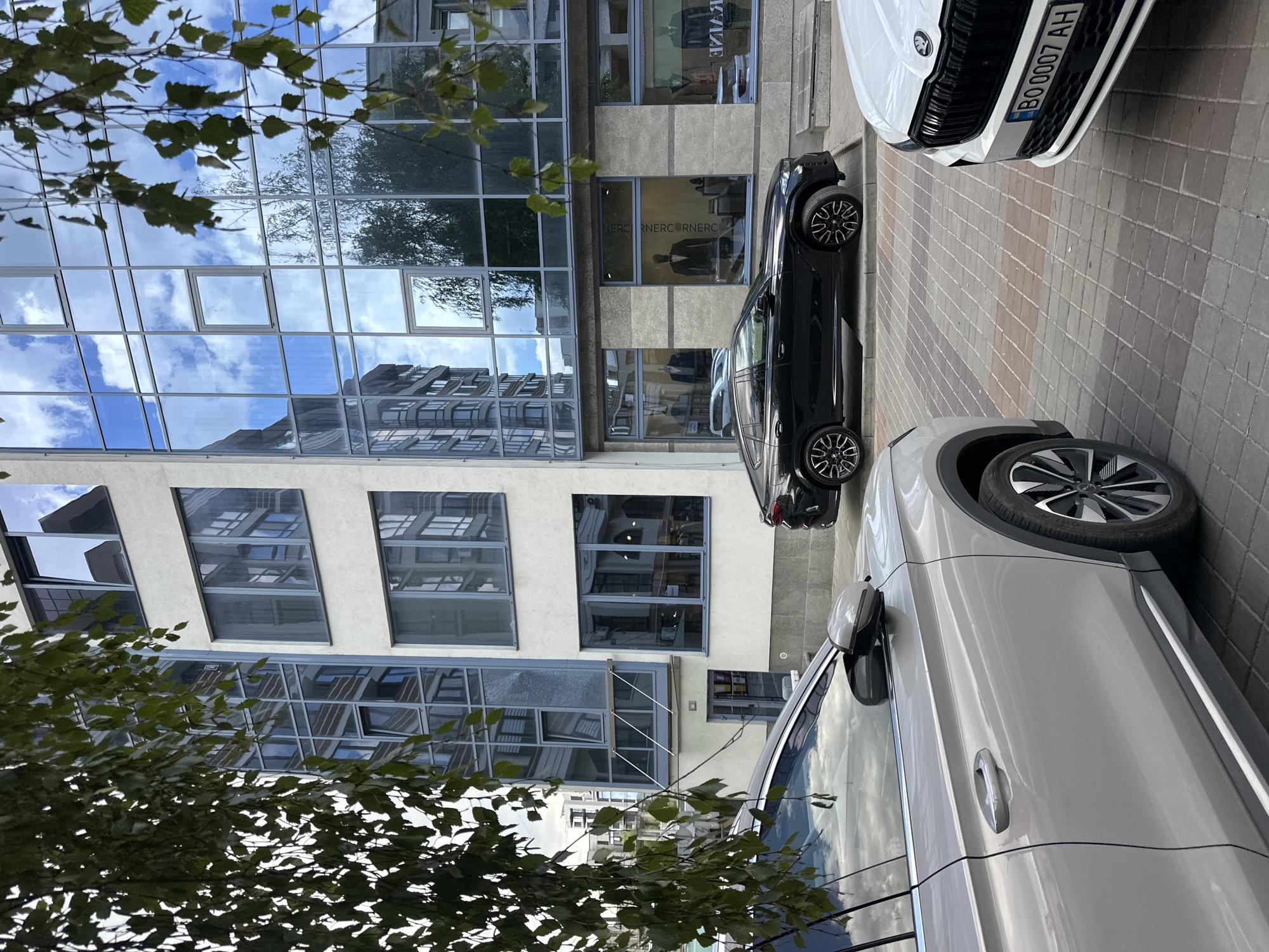 In Ternopil, Ukraine, a city scene unfolds with a modern building as the backdrop. The building features a facade with large glass windows that reflect the sky, clouds, and surrounding architecture, indicating an urban setting. In the foreground, three cars are parked on a patterned brick sidewalk. The closest car, on the left, is a light-colored SUV, partially visible, showing its side profile, door handle, and wheel. To its right is a black coupe SUV, also parked. Further to the right, the front of a white SUV is visible, displaying its license plate which reads "BO 0007 AH" and the Skoda logo on its grille. The ground is paved with light-colored and reddish-brown bricks arranged in a grid pattern. Lush green foliage hangs from the upper left and right edges of the frame, suggesting the presence of trees or bushes adjacent to the scene. The building's lower level has storefronts with mannequins displaying suits, implying a commercial area.