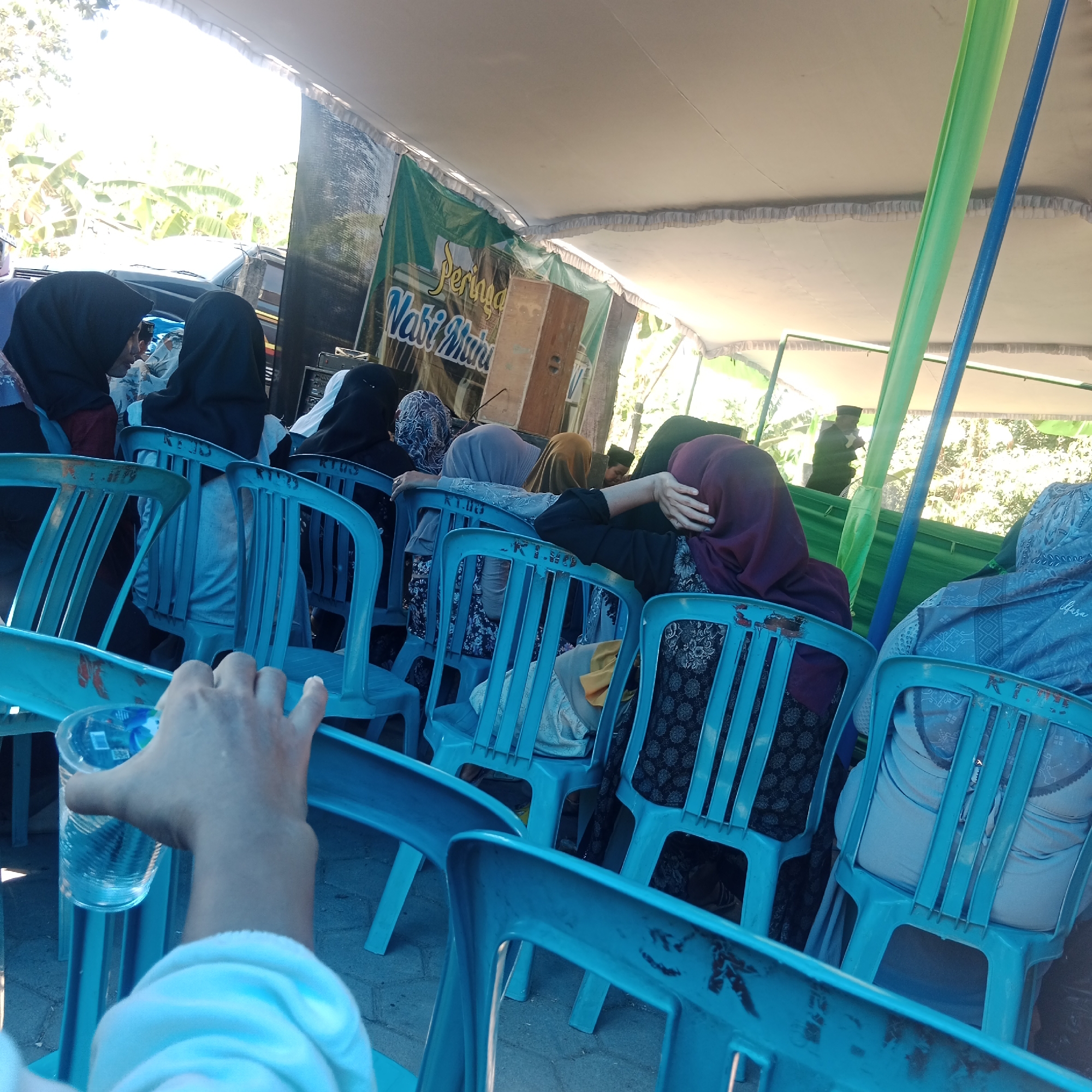 The image depicts an outdoor gathering under a white tent. Several individuals, predominantly women wearing hijabs, are seated on blue plastic chairs, facing towards a banner at the front that reads "Peringatan Maulid Nabi Muhammad" (Commemoration of the Prophet Muhammad's Birthday). The setting appears to be in Tegowanu Kulon, Indonesia, suggested by the banner's text and common use of such tents for community events in the region. The lighting indicates it is daytime, with sunlight filtering through the tent and surrounding foliage. One person in the foreground is holding a clear plastic cup with water, their arm and hand prominently in view. The background shows trees and a vehicle, hinting at a semi-rural or suburban location. The overall atmosphere suggests a religious or cultural event.