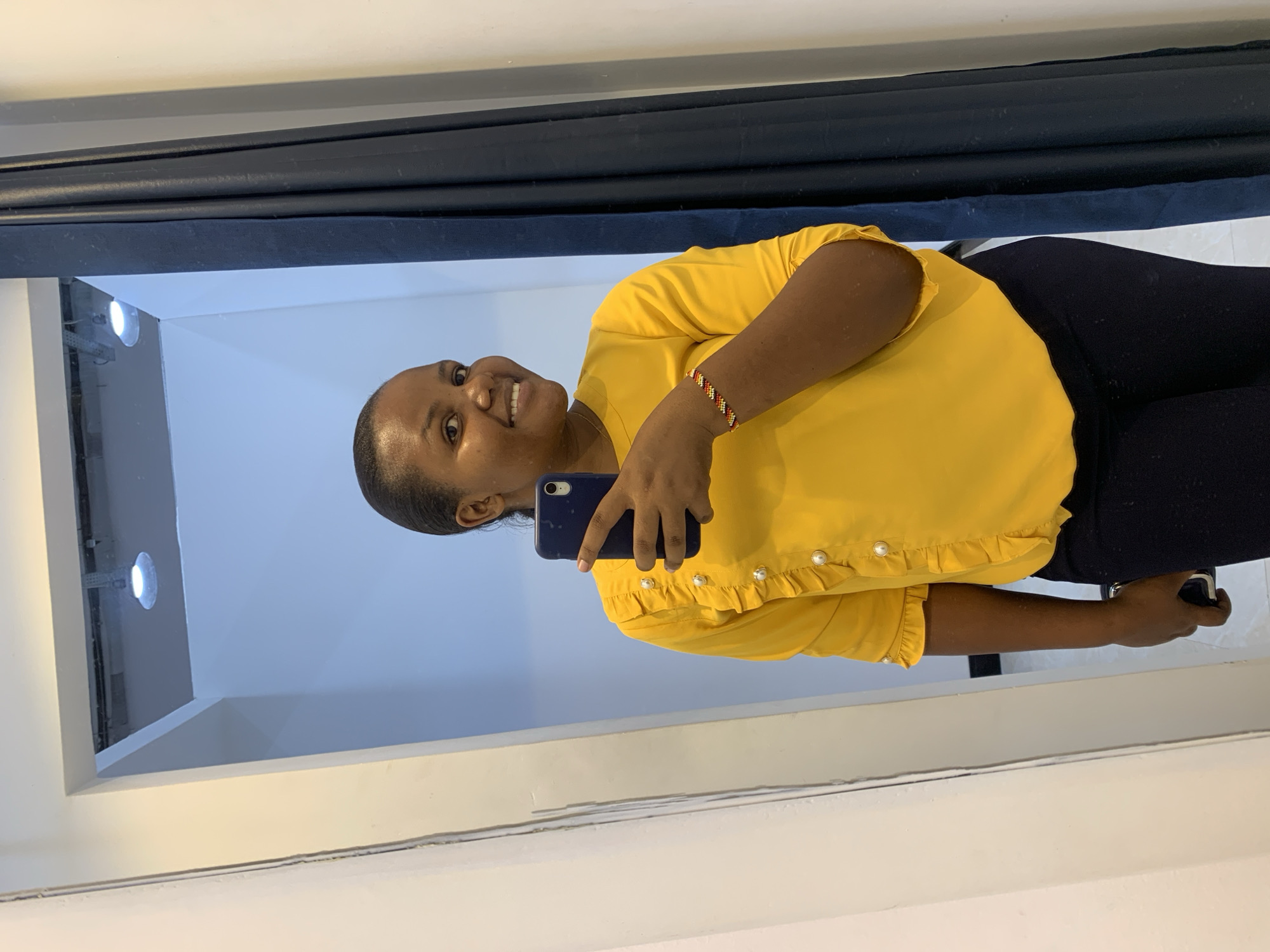 A woman in a yellow blouse and black pants takes a selfie with a blue smartphone in a room in Dar es-Salaam, Tanzania. She has a beaded bracelet on her left wrist. The room appears to be small, with a white wall and a blue curtain. The woman is smiling at the camera.