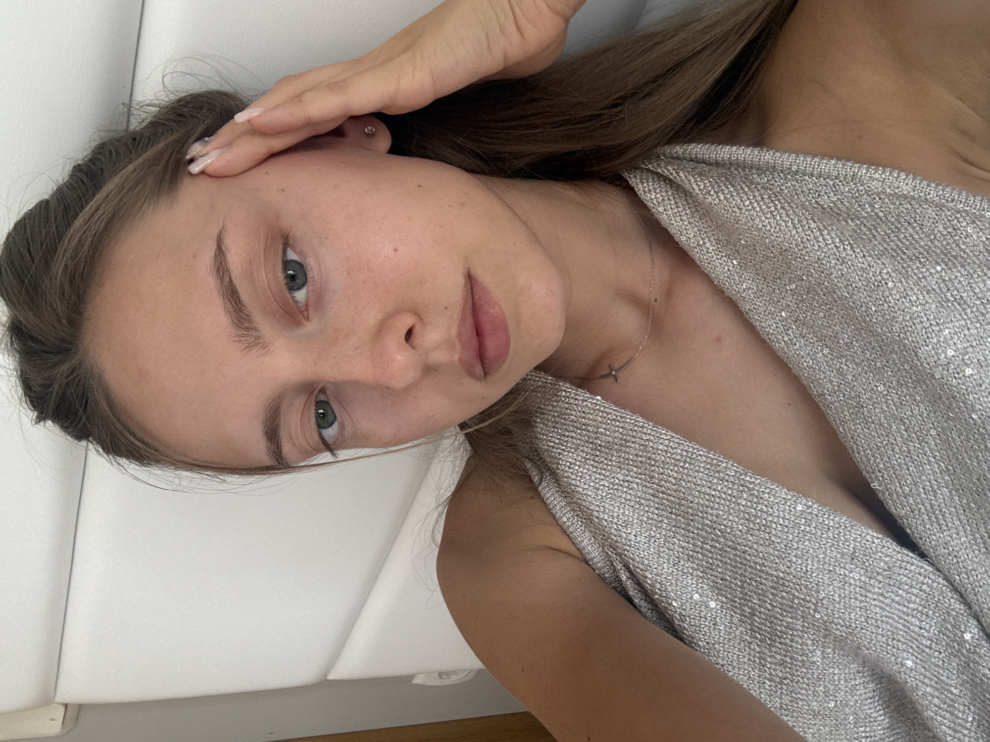 A young woman with light brown hair is lying on her side in bed, facing the camera. She is wearing a silver sequined top. Her right hand is raised to her forehead, and her eyes are looking at the camera. The background is a white padded headboard of a bed. The image does not provide any information about the location in Sozopol, Bulgaria.
