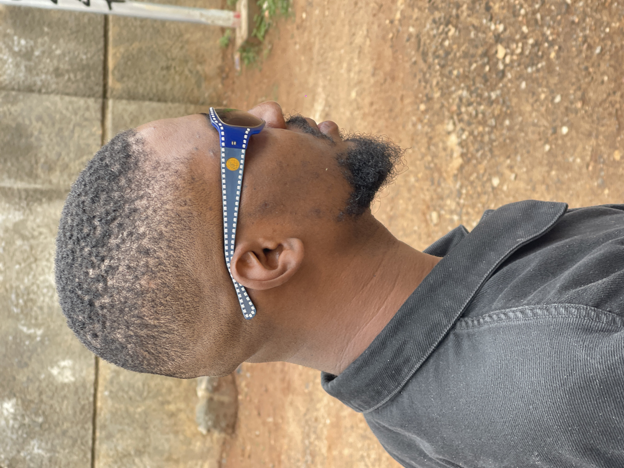 A man is shown from the side, wearing sunglasses and a dark shirt. He has short, textured hair and a goatee. The background consists of a textured wall and a dirt path with small rocks. The lighting suggests it is daytime. The scene appears to be outdoors in an urban or semi-urban environment.