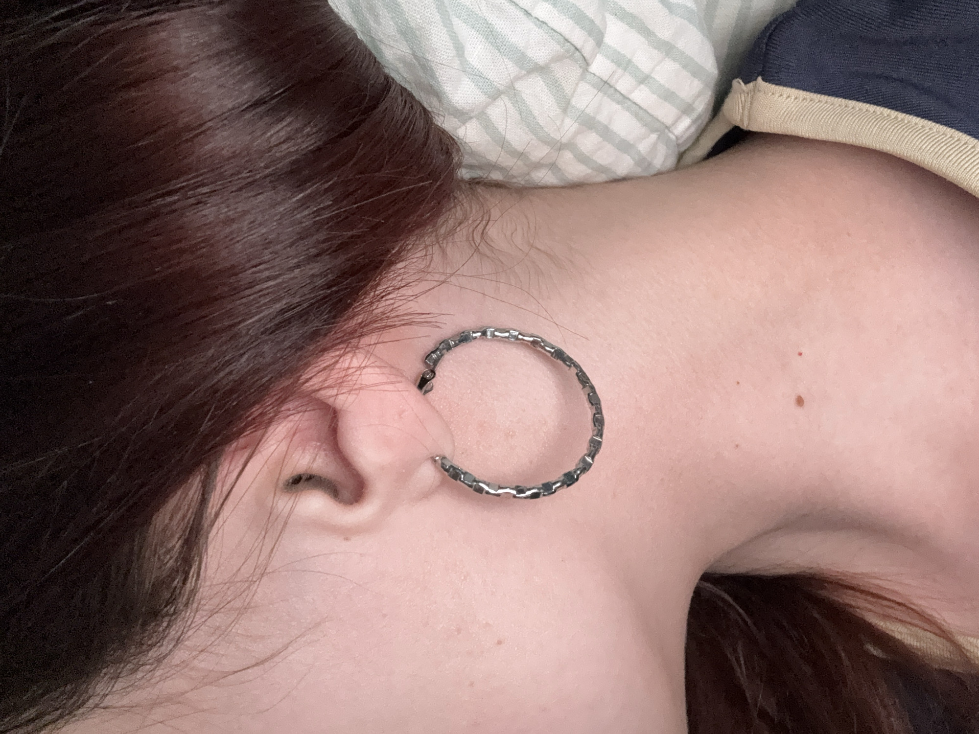 A close-up shot of a person's head and upper body lying on a pillow. The person has dark brown hair and pale skin. They are wearing a silver hoop earring in their left ear. The pillow has a striped pattern. A mole is visible on the person's neck. The image does not provide information to confirm location in Прикамский, Russia.