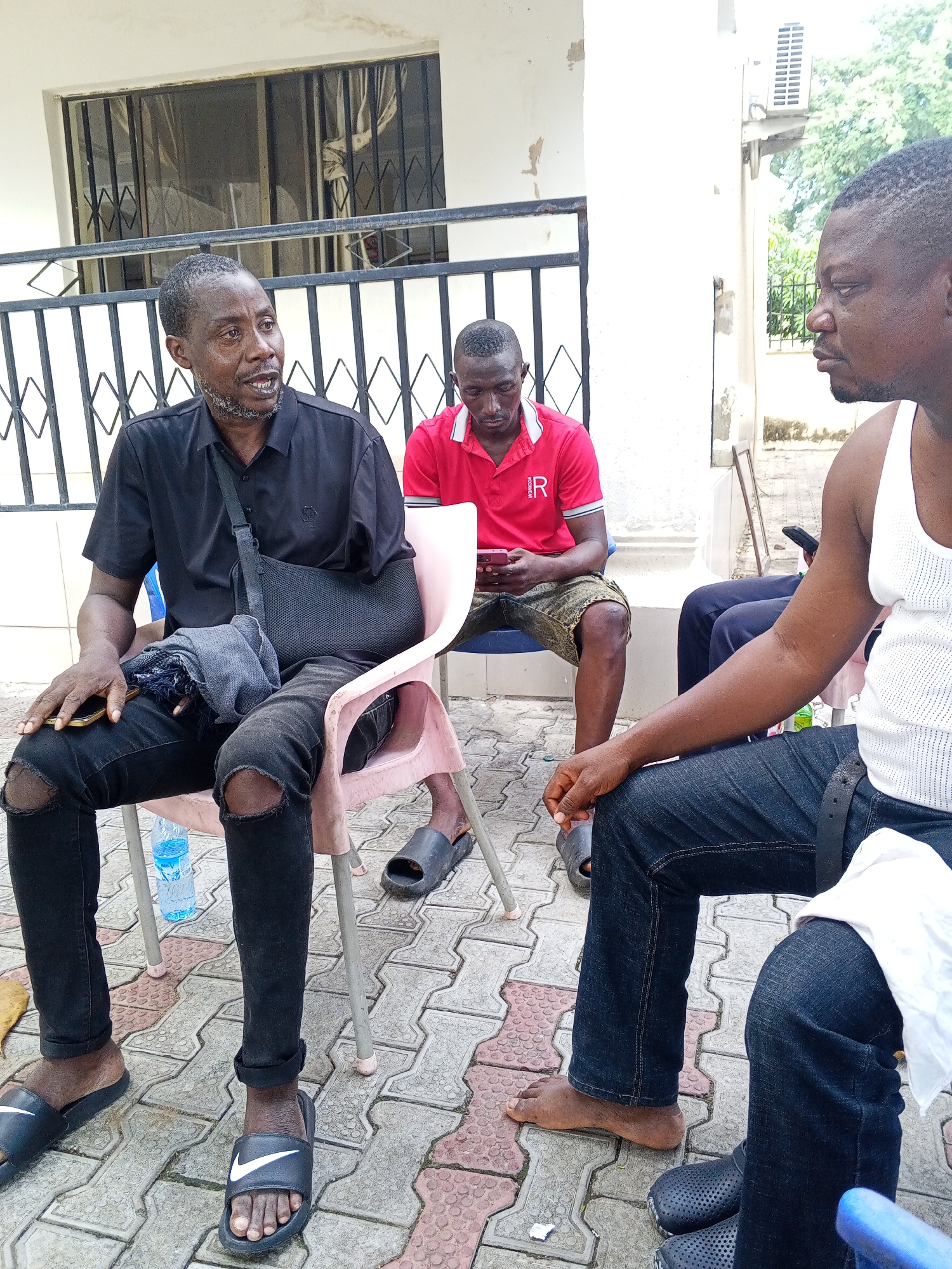 The image displays three men sitting outdoors on a paved area with interlocking bricks. The central figure, seated on a pink plastic chair, is a man wearing a black short-sleeved collared shirt and ripped black jeans. His left arm is in a black sling, and he holds a dark mobile phone in his right hand. He is speaking, looking towards the right side of the frame, and wears black Nike slides.
To his right, partially visible, sits a man in a red polo shirt with the letter "R" on it, and patterned shorts. He is looking down at a mobile phone in his lap. On the far right, another man in a white tank top and dark jeans is seated, looking towards the man in the black shirt. He has black perforated slides on one foot and is barefoot on the other. A glimpse of another person's legs can be seen behind him.
In the background, a white building features a window with black security bars and an external air conditioning unit. A black metal railing runs horizontally behind the seated men. The scene is brightly lit by natural light, indicating daytime. The casual attire suggests a warm climate. The men appear to be engaged in a conversation or a casual gathering.