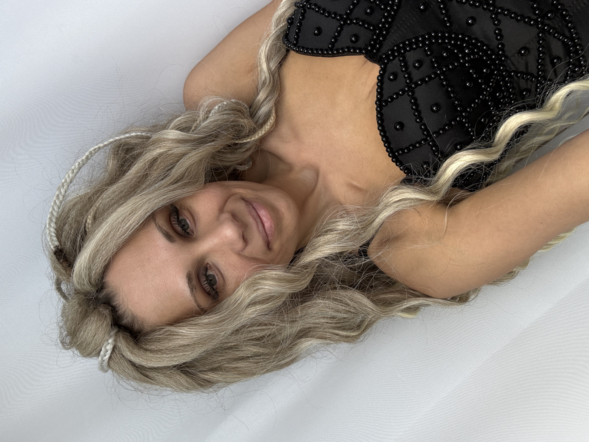 A woman with long, wavy, blonde hair is lying on a white surface. She is wearing a black top with black embellishments. Some strands of her hair are braided. The location is unknown.