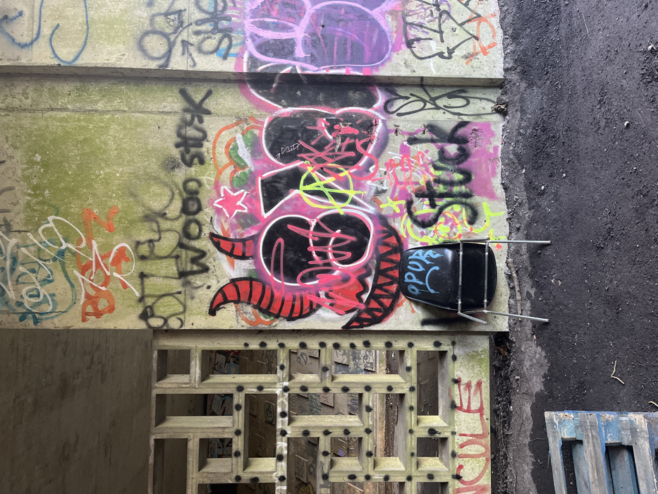 Graffiti covers a concrete wall, featuring bold black and red designs with pink and green accents. An overturned black chair with "OPOR" scrawled on its back sits beside the graffiti. Below, a patterned concrete block wall with small openings reveals a glimpse into a darker space beyond. To the right, dark asphalt and a blue wooden pallet add texture to the scene.