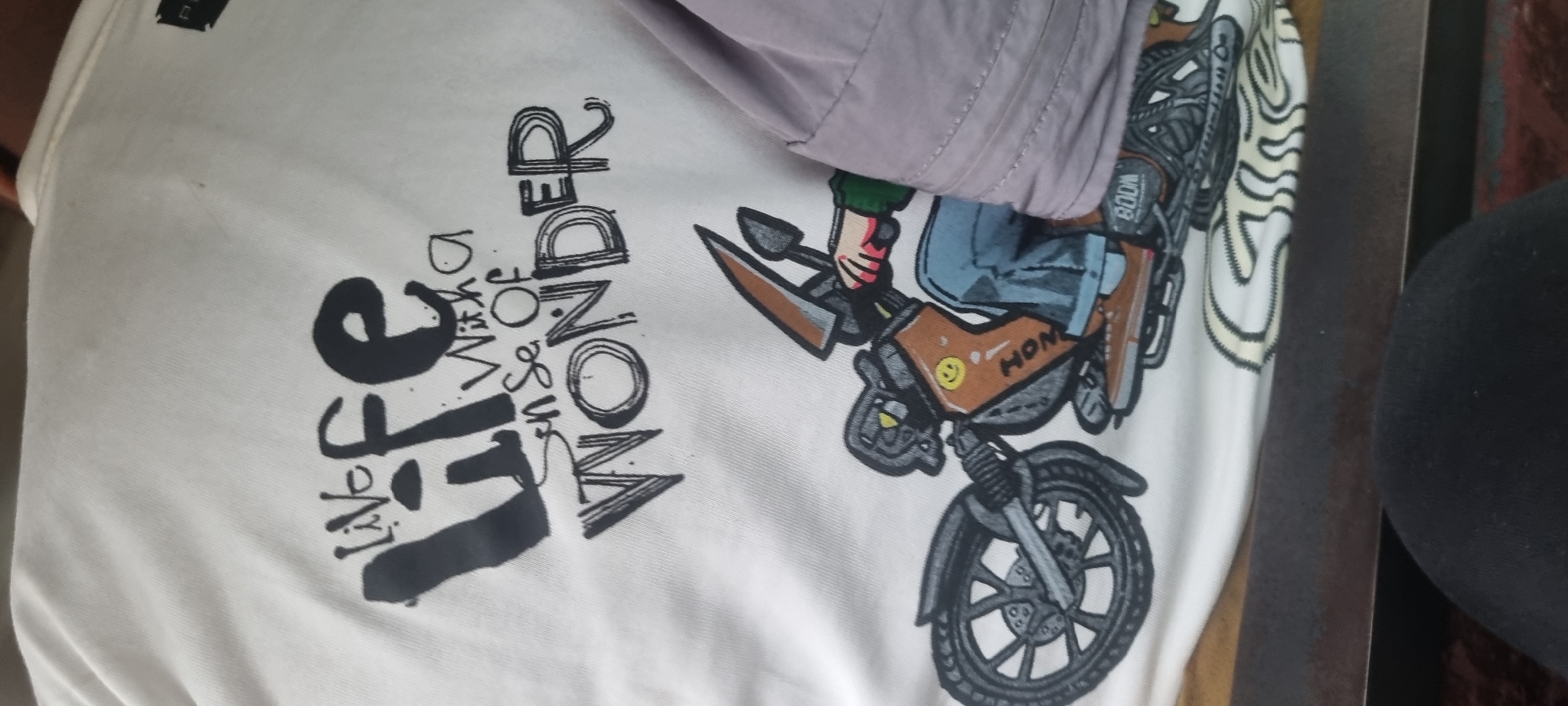 The image is a close-up shot of a white t-shirt with a graphic print. The graphic features text and an illustration of a person riding a motorcycle. The text reads "Live Life With A Sense Of WONDER." Below the text is a cartoonish drawing of a motorcycle with a rider. The rider is wearing blue jeans and brown shoes. The motorcycle has details like a smiley face, the word "HONM" on the side, and "BOOM" on the rear. The t-shirt is made of a ribbed fabric. The overall tone of the image is casual and artistic. The scene is indoors, as indicated by the artificial lighting and the presence of a wooden surface below the t-shirt. The specific location in Agege, Nigeria is not discernible from the image itself.