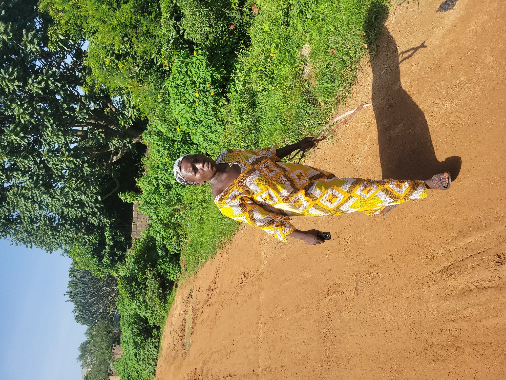 A woman walks down a dirt road in Jos, Nigeria. She is wearing a yellow and white patterned dress and a headscarf. She is holding a mobile phone in her left hand and a stick in her right hand. The road is dusty and uneven, with puddles of water in places. There are trees and green bushes on either side of the road. The sky is blue and clear. The sun is shining, casting a long shadow from the woman on the ground.