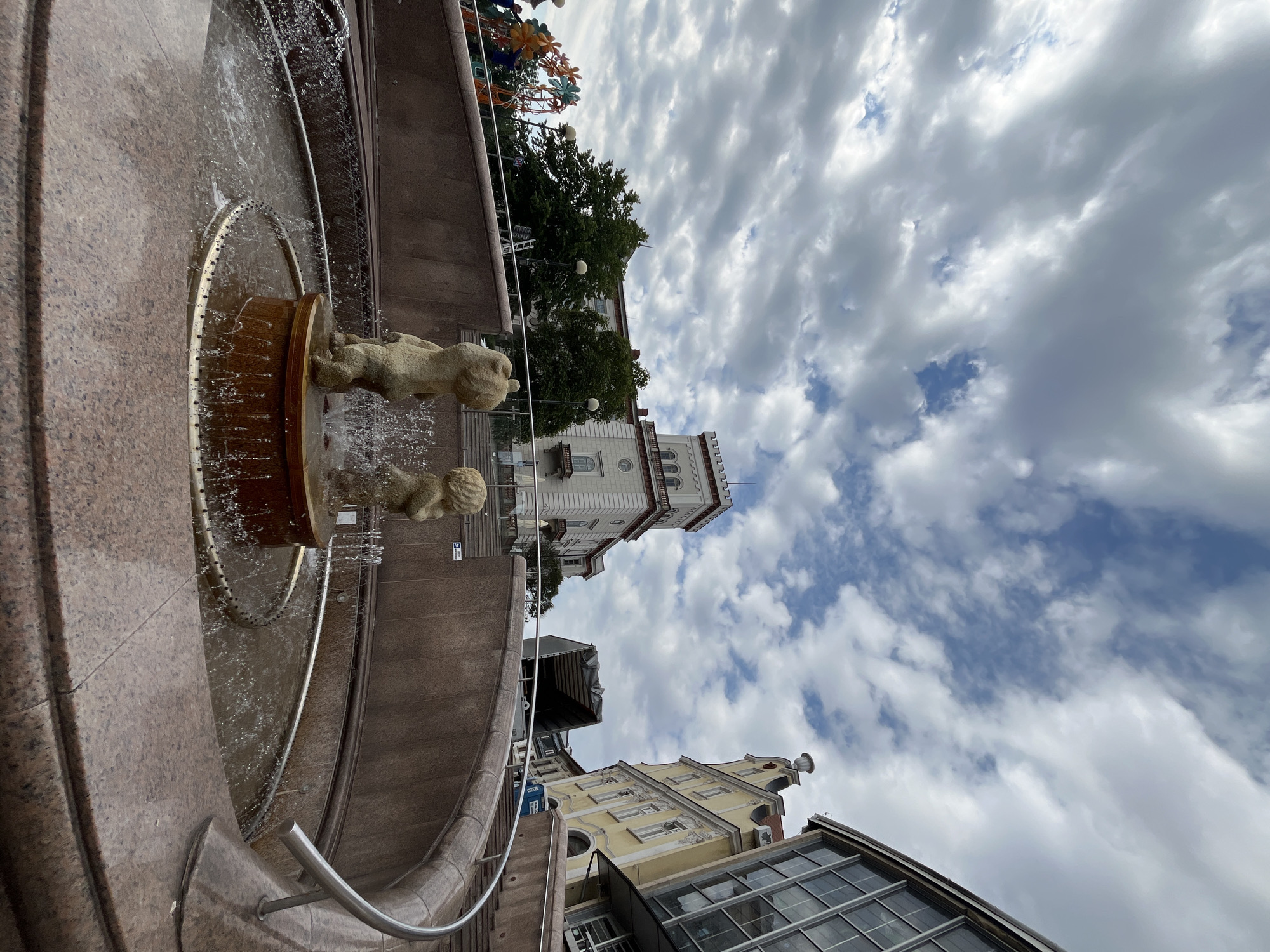 An eye-level, wide-angle shot captures a tiered fountain in the foreground, with two stone figures of children appearing to play in its waters. The fountain is made of dark red granite and is surrounded by a circular stone wall. In the background, a wide set of stairs leads up to a plaza with several buildings. To the left of the stairs, a playground with colorful structures is visible. The buildings in the background include a light-colored building with a tower and crenellations, a yellow building with ornate architectural details, and a modern glass building. The sky above is filled with dramatic, grey and white clouds, suggesting an overcast day.