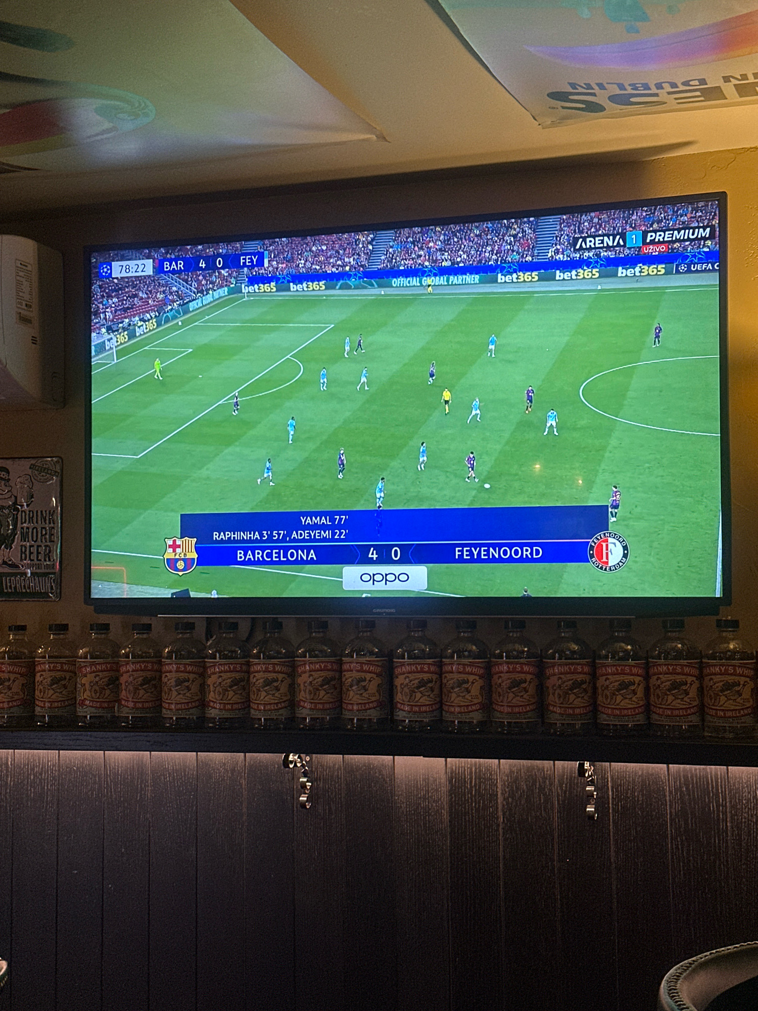 The image shows a television screen displaying a live broadcast of a football match. The score indicates Barcelona is leading Feyenoord 4-0. The match is part of the UEFA Europa League, as suggested by the logos and sponsorship banners visible. Below the television, a row of identical bottles, likely alcoholic beverages with labels that read "SHANKY'S WHIP MADE IN IRELAND," are displayed on a shelf. The background behind the bottles appears to be a dark wooden paneling. The scene is indoors, possibly in a bar or a private viewing area, given the sporting broadcast and the arrangement of the bottles. No specific location in Belgrade, Serbia can be identified from the visual information presented.