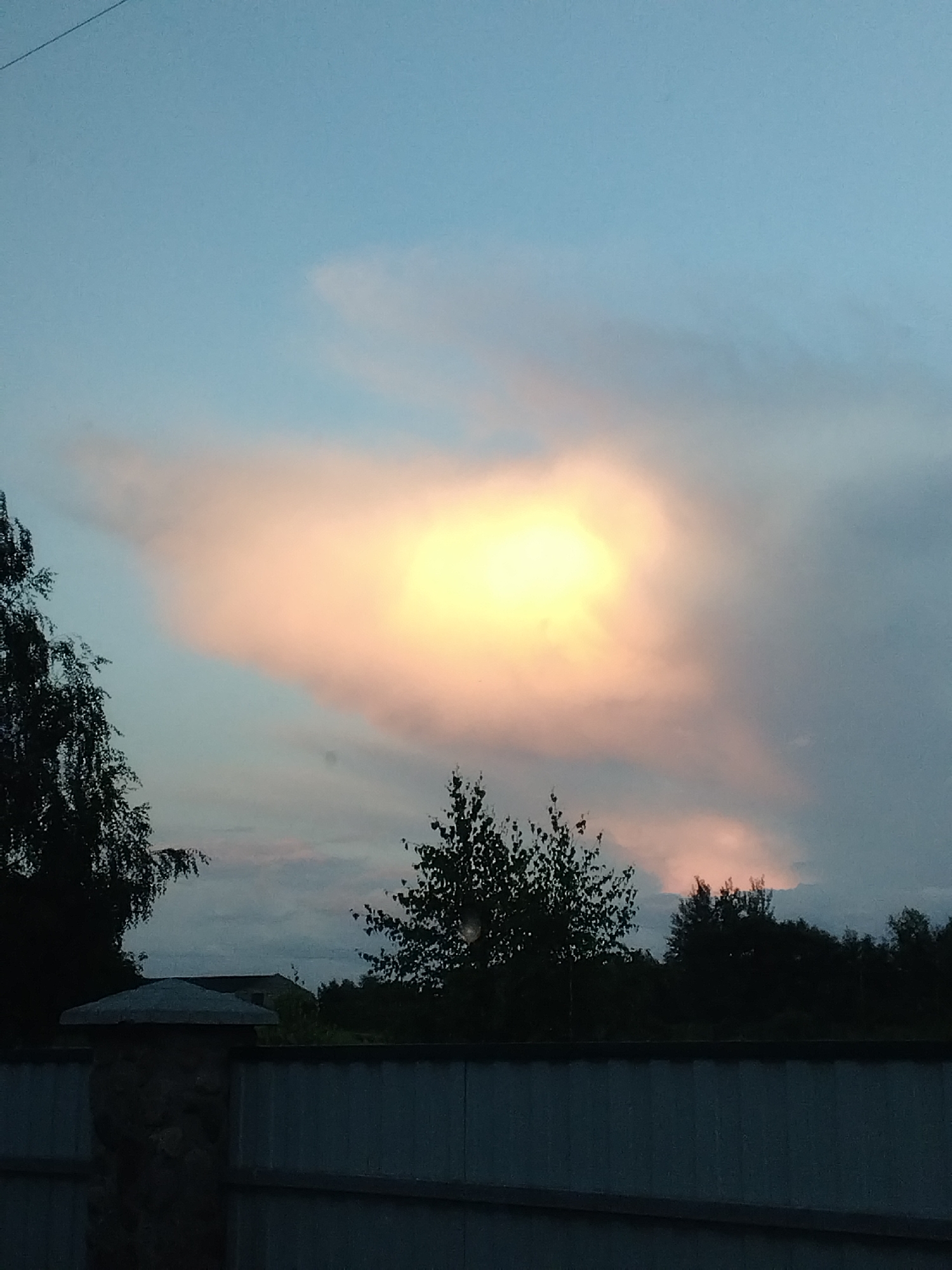 This is a horizontal, medium shot, taken outdoors at dusk. The sky is a soft blue, transitioning to darker grey, with a bright, luminous cloud formation in the center, tinged with pink and yellow hues. Silhouetted against the sky are the dark, dense shapes of trees. In the foreground, a weathered, grey metal fence runs horizontally across the bottom of the frame, with a stone pillar on the left. The location appears to be rural, possibly within or near the Свіслацкі сельскі Савет in Belarus. No people or animals are visible. The overall impression is of a peaceful, natural scene at the end of the day.